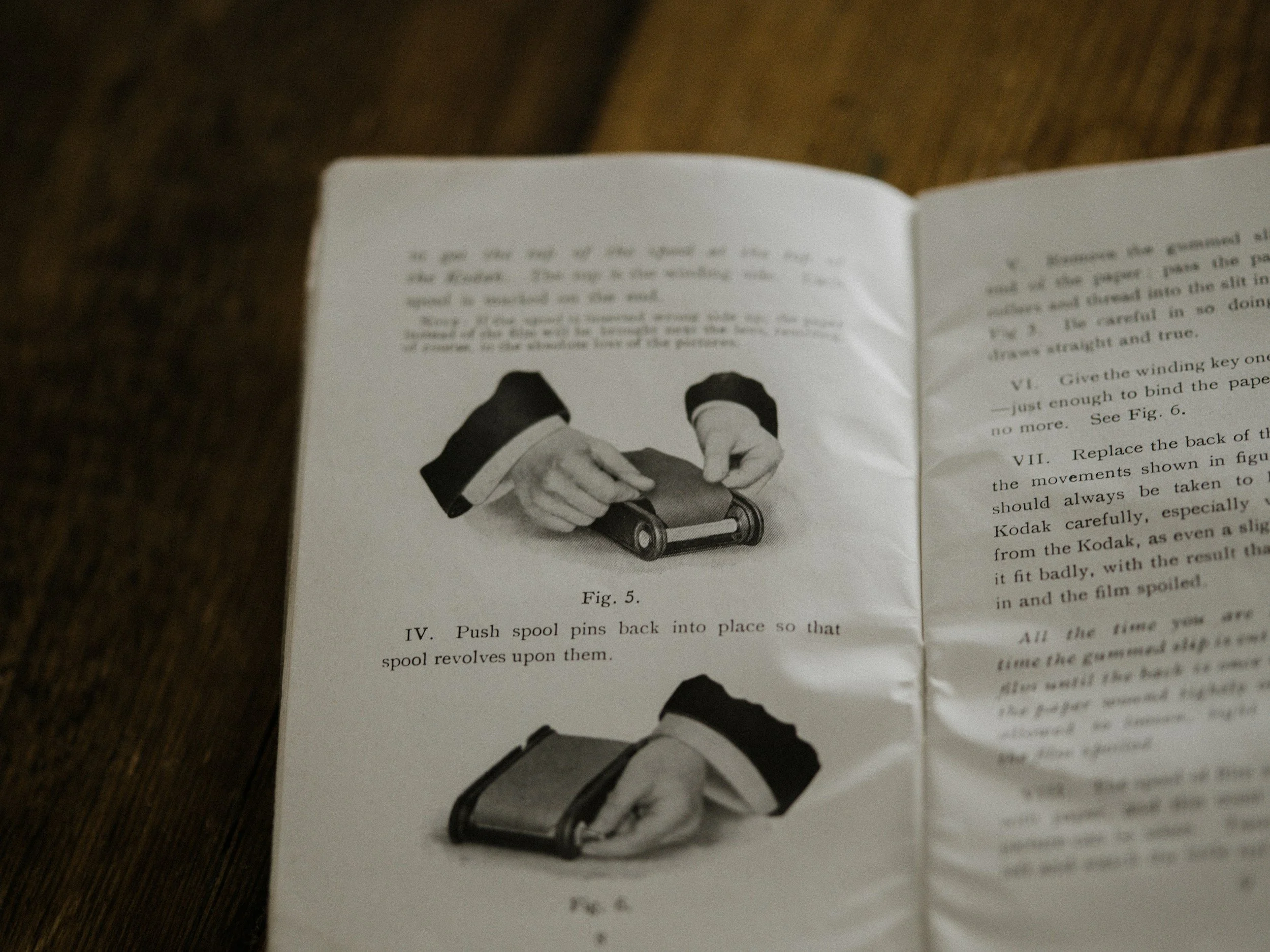 A blurry photograph of an open instruction manual or booklet with black-and-white illustrations and text, placed on a wooden surface.