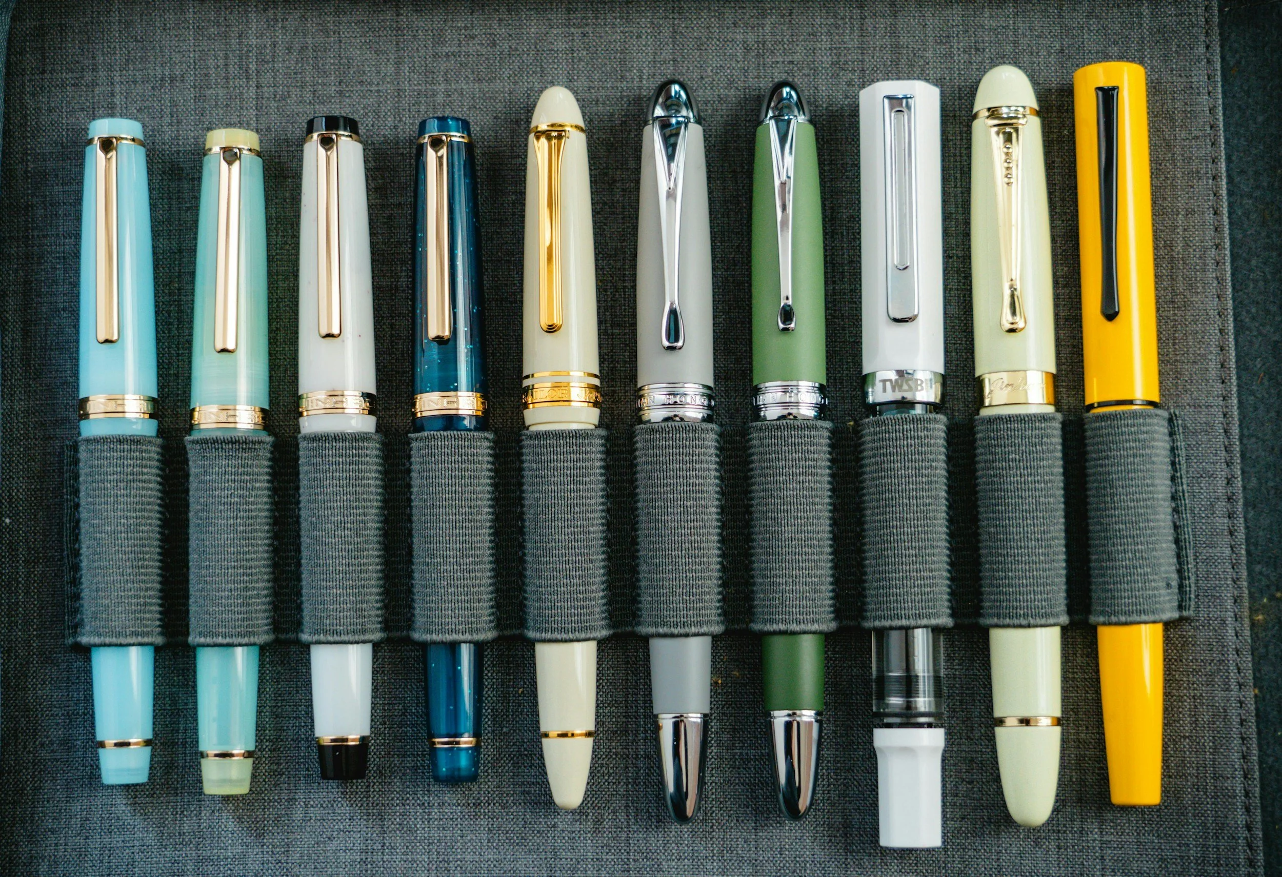 A collection of colorful fountain pens arranged side by side on a dark fabric surface.