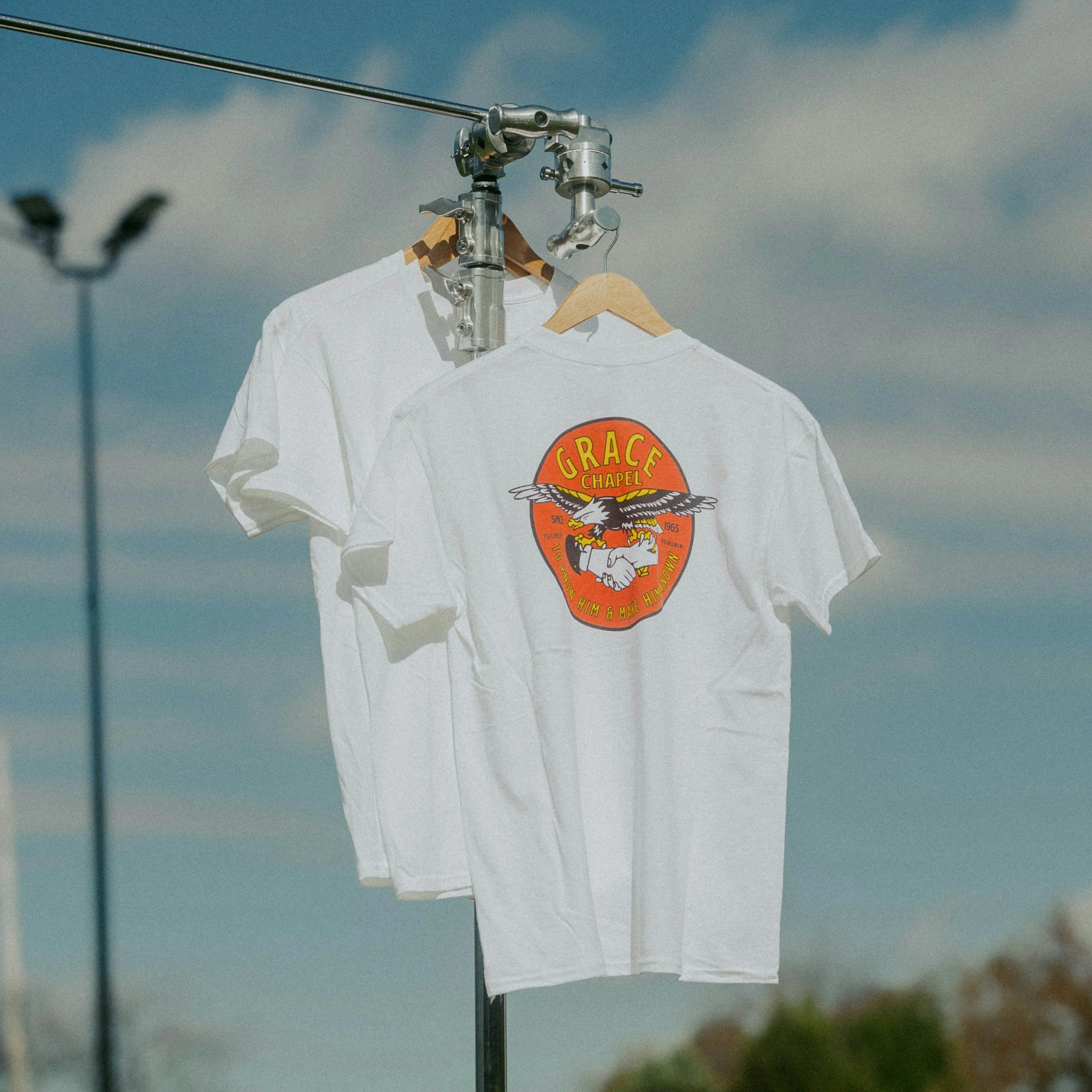 A white T-shirt with a colorful graphic and the text "Grace Chapel" hung on a wooden hanger from a metal clothing rack against a cloudy sky.