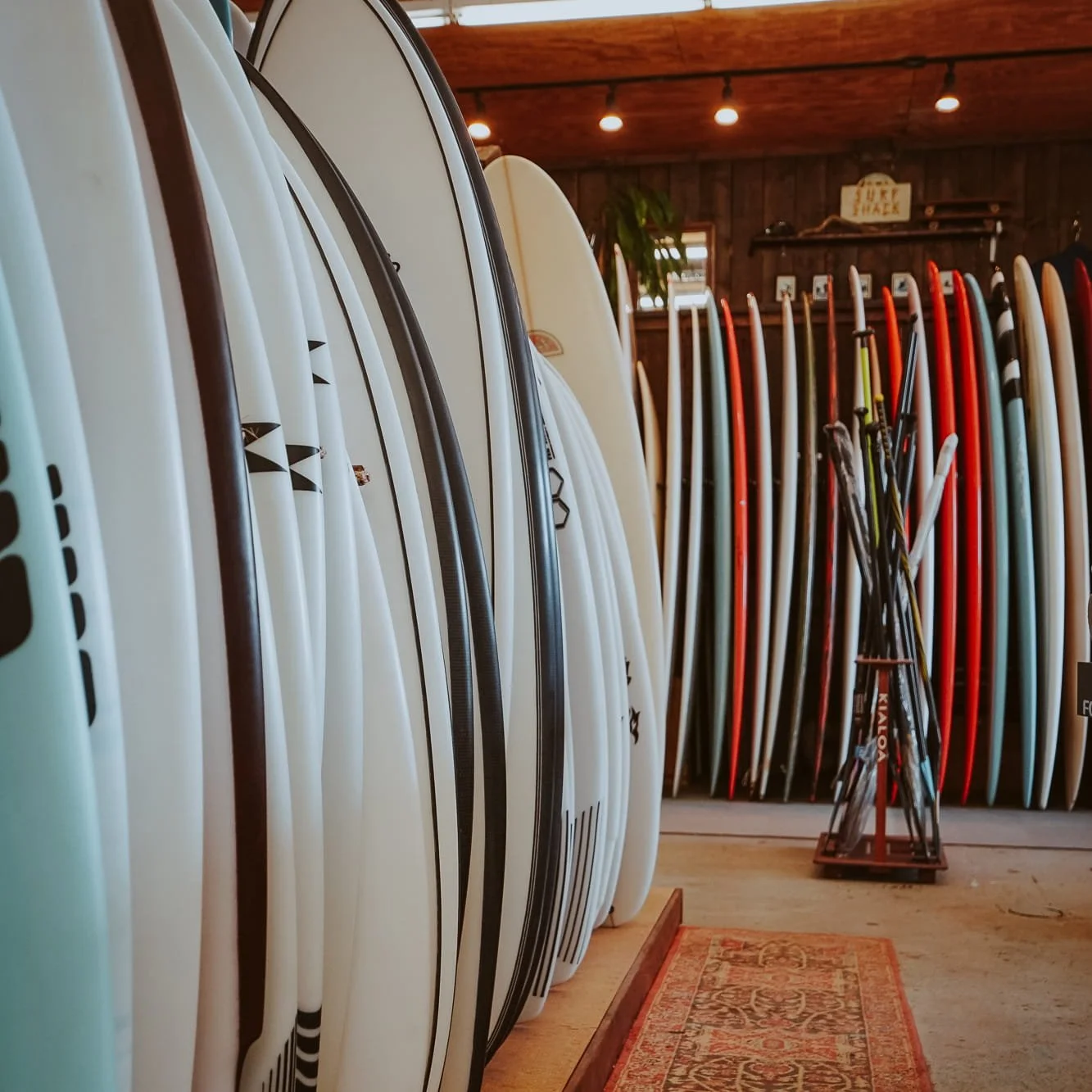 Rows of surfboards and paddleboards in a shop with wooden walls and ceiling, with some paddles in a stand in the center.