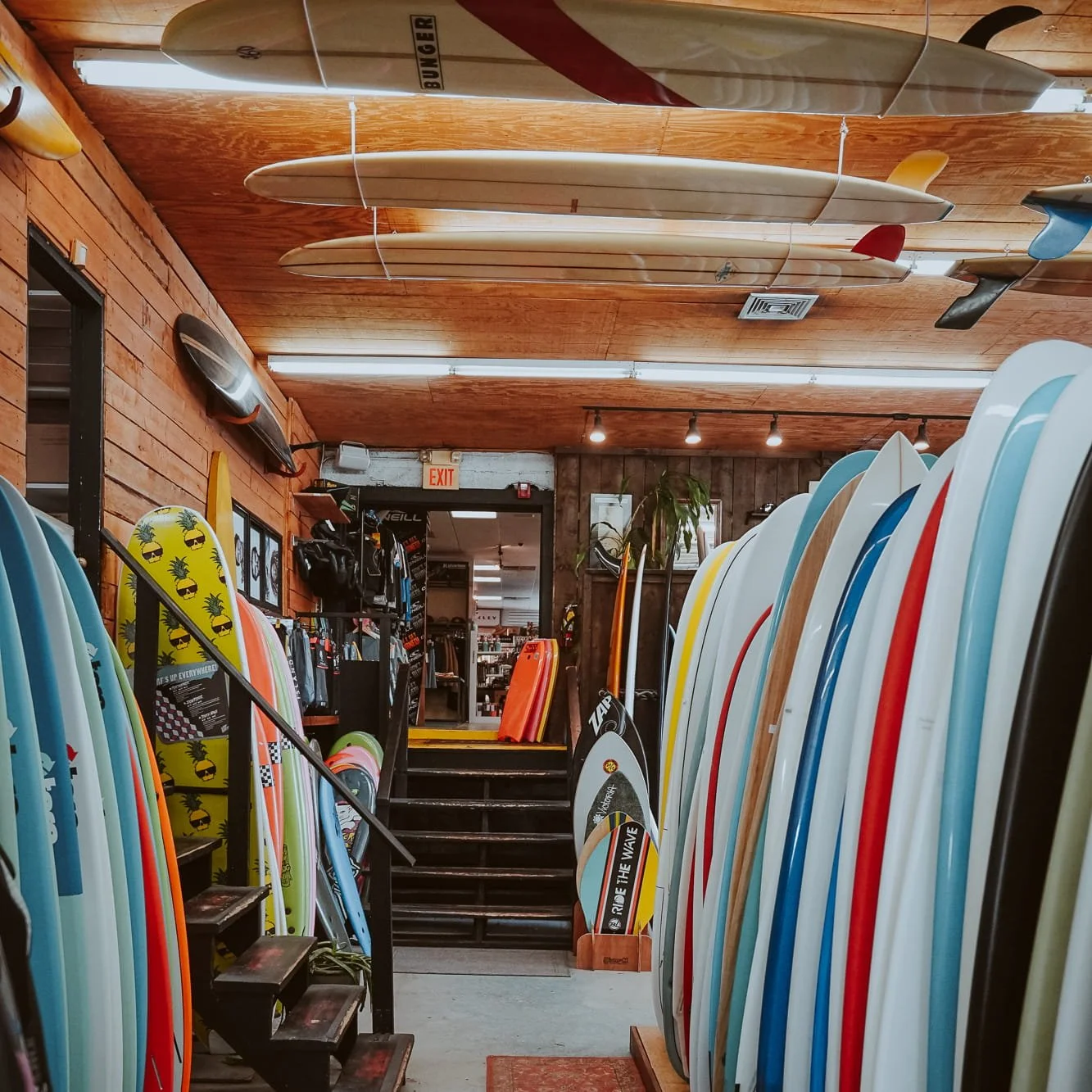 Store interior with surfboards of various colors and sizes displayed vertically on shelves, hanging surfboards on the ceiling, and skateboards and apparel on the back wall.