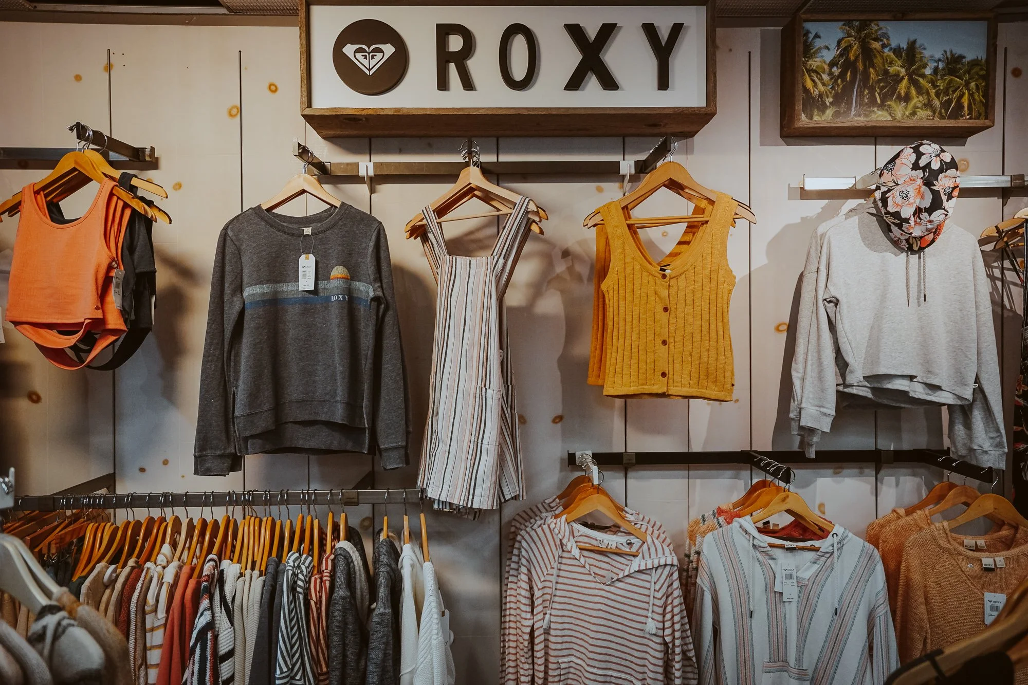 Clothing display at a shop with a sign reading 'ROXY' on the wall. Various shirts, dresses, and jackets on hangers, including a gray sweatshirt, striped dresses, and a yellow vest, with a floral scarf on a mannequin.