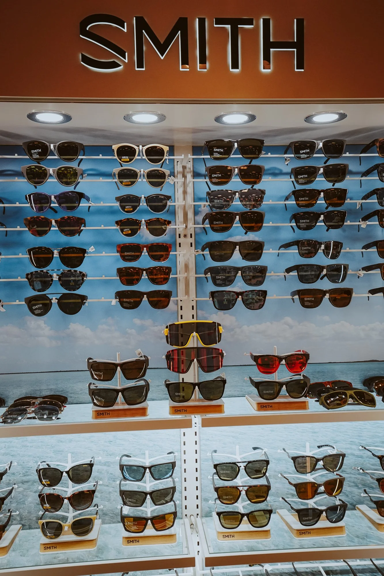 A display of various sunglasses on shelves with a scenic beach background and a sky with clouds.