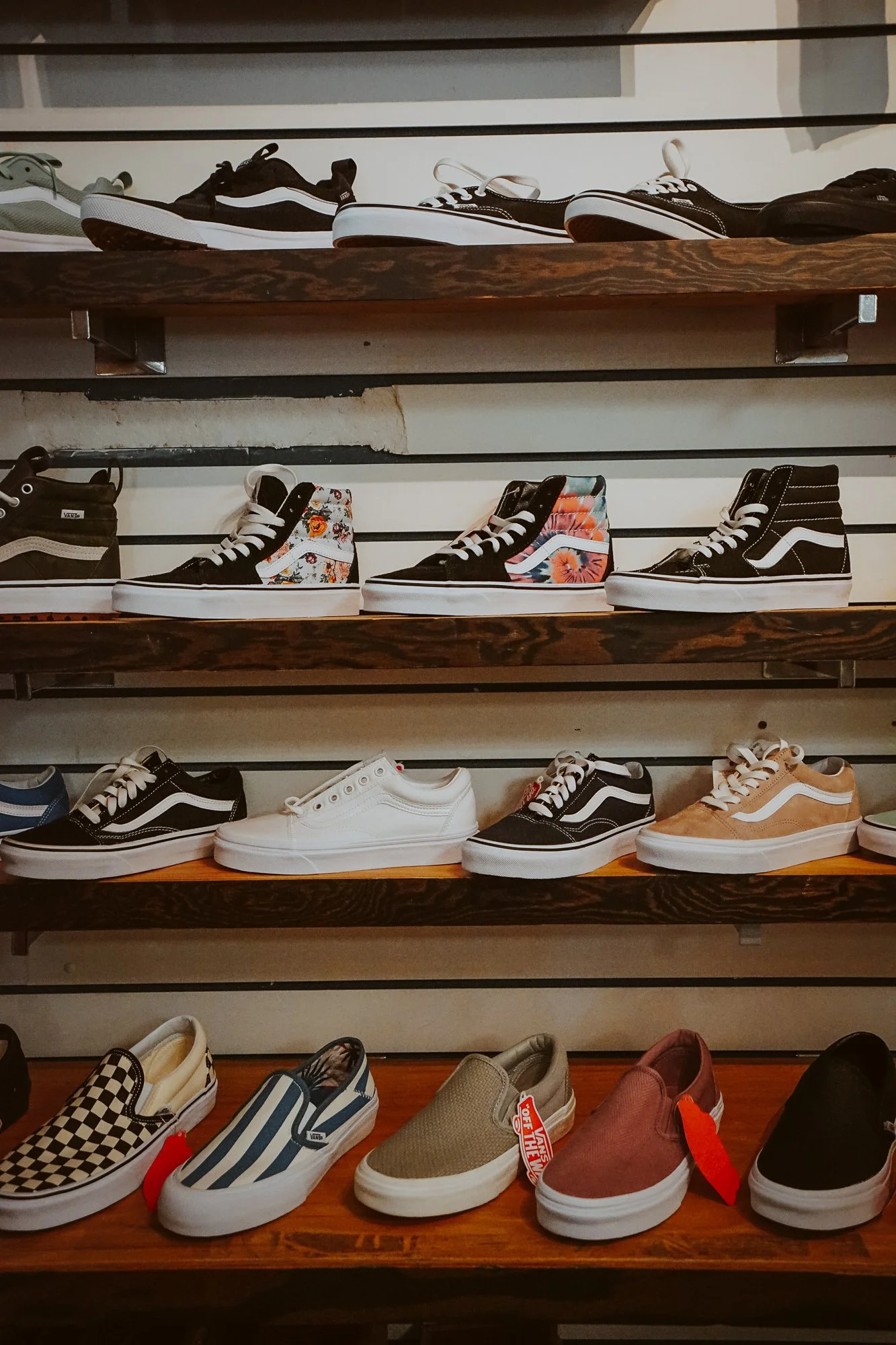 Display of various Vans sneakers on wooden shelves, including checkered, striped, floral, solid colors, and high-tops.