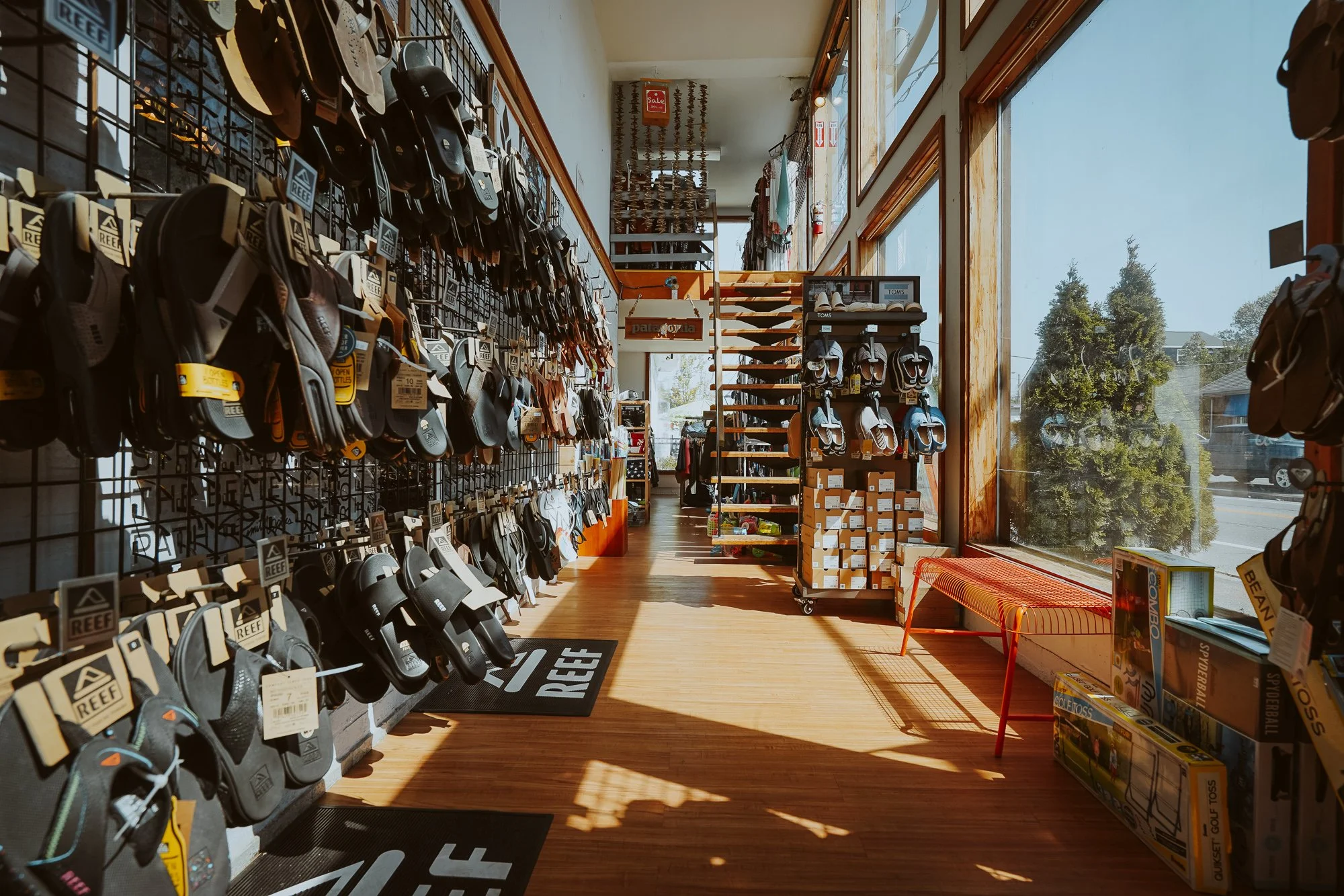 Interior of a shoe store with shelves holding sandals and flip-flops, a staircase in the background, large windows letting in natural light, and a bench near the window.