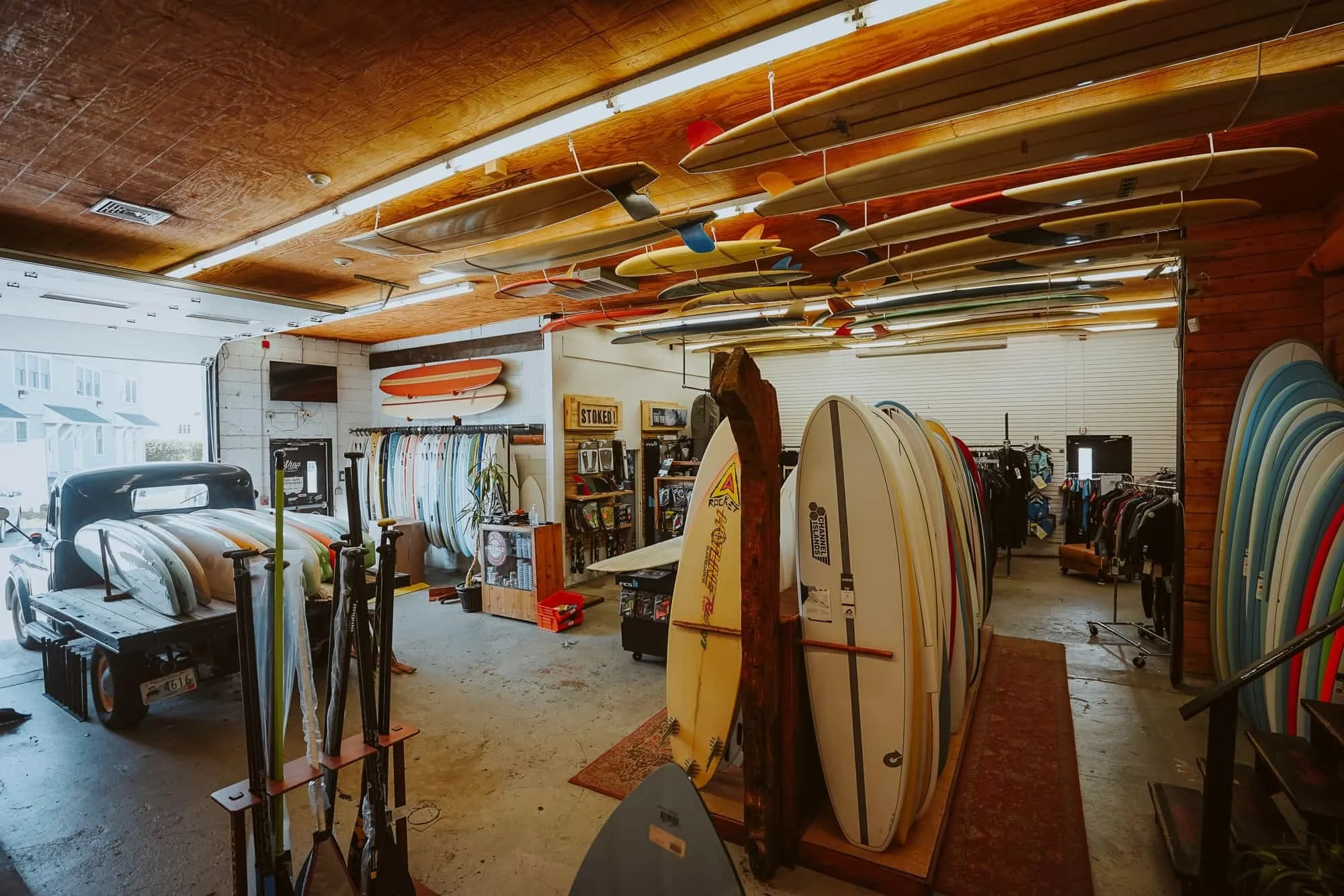 Indoor surf shop displaying surfboards hanging from the ceiling, standing surfboards and fins arranged in racks, and a vintage truck with surfboards in the foreground, with clothing and accessories on shelves and racks inside.