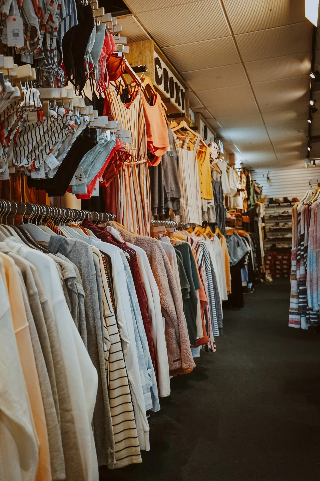Clothing store aisle with various shirts, jackets, and pants hanging on racks, and a sign with the logo of the store in the background.