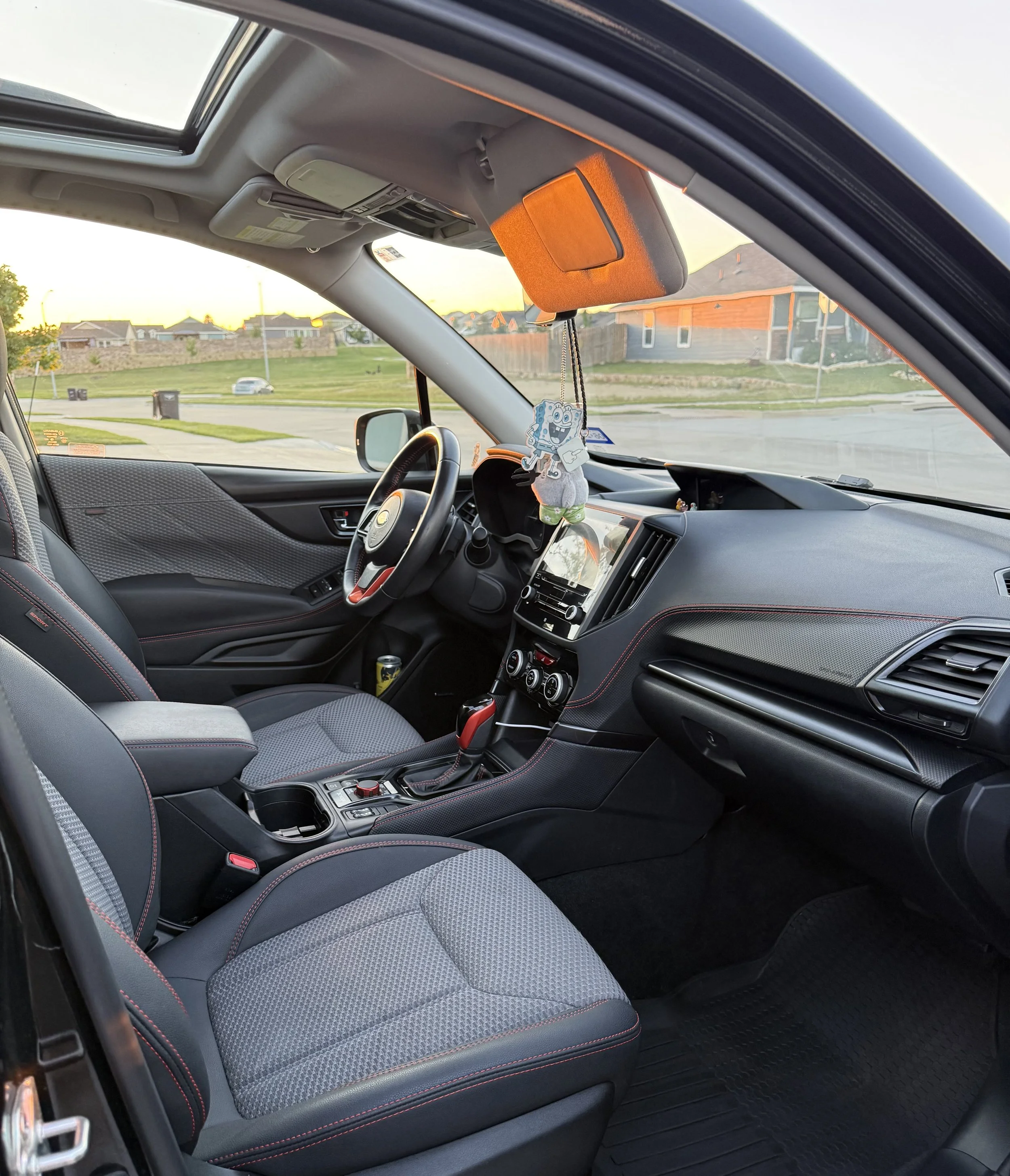 The interior of a modern car showing the front seats, dashboard, steering wheel, and part of the windshield with a view of a suburban neighborhood and sunset outside.