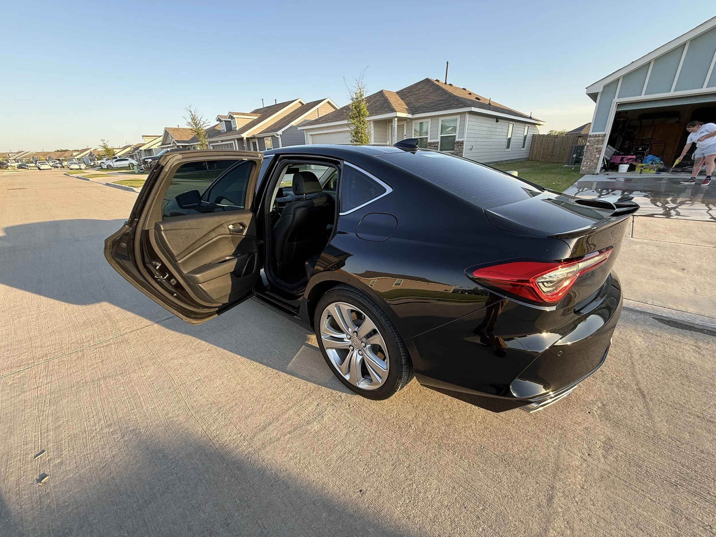 A black modern sedan car with open rear passenger door parked on a suburban street, with houses, trees, and a garage in the background.