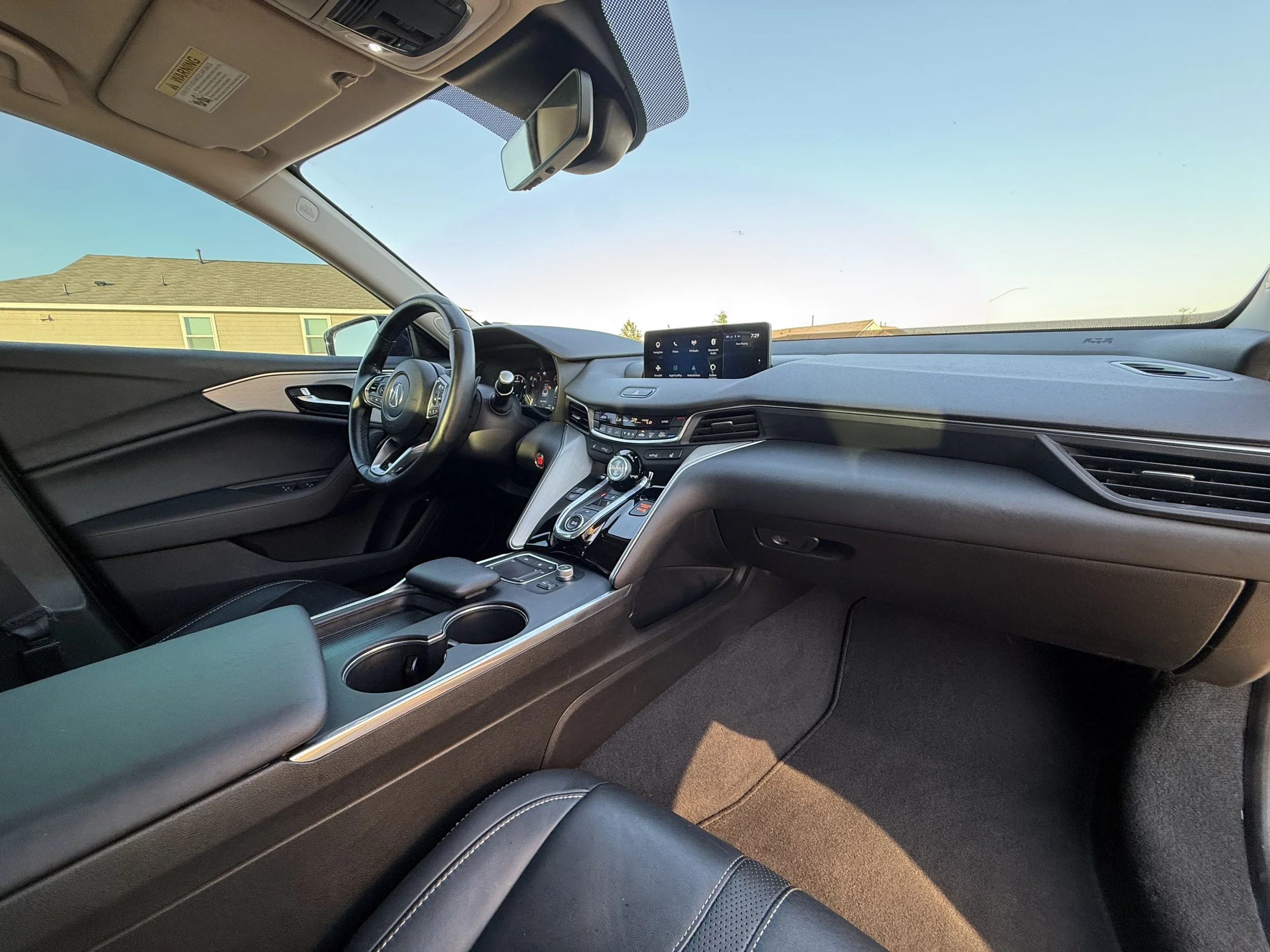 Interior of a modern car showing the driver's seat, dashboard, center console with climate controls, digital display, gear shifter, and a rearview mirror, with a view of the clear sky and suburban rooftops through the windshield.