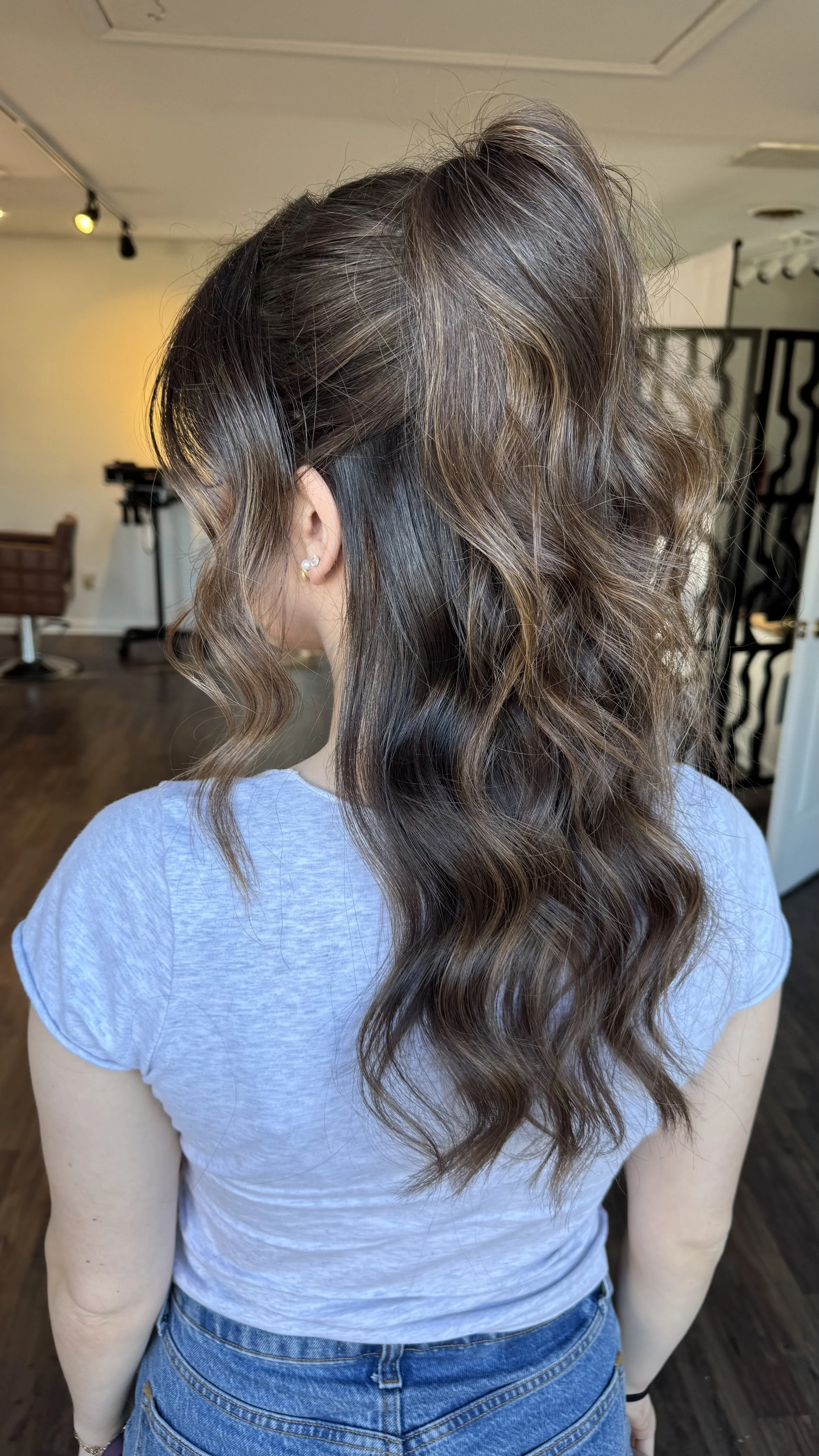 A woman with long, wavy brown hair with blonde highlights, wearing a light gray T-shirt and blue jeans, is standing in a salon or studio.