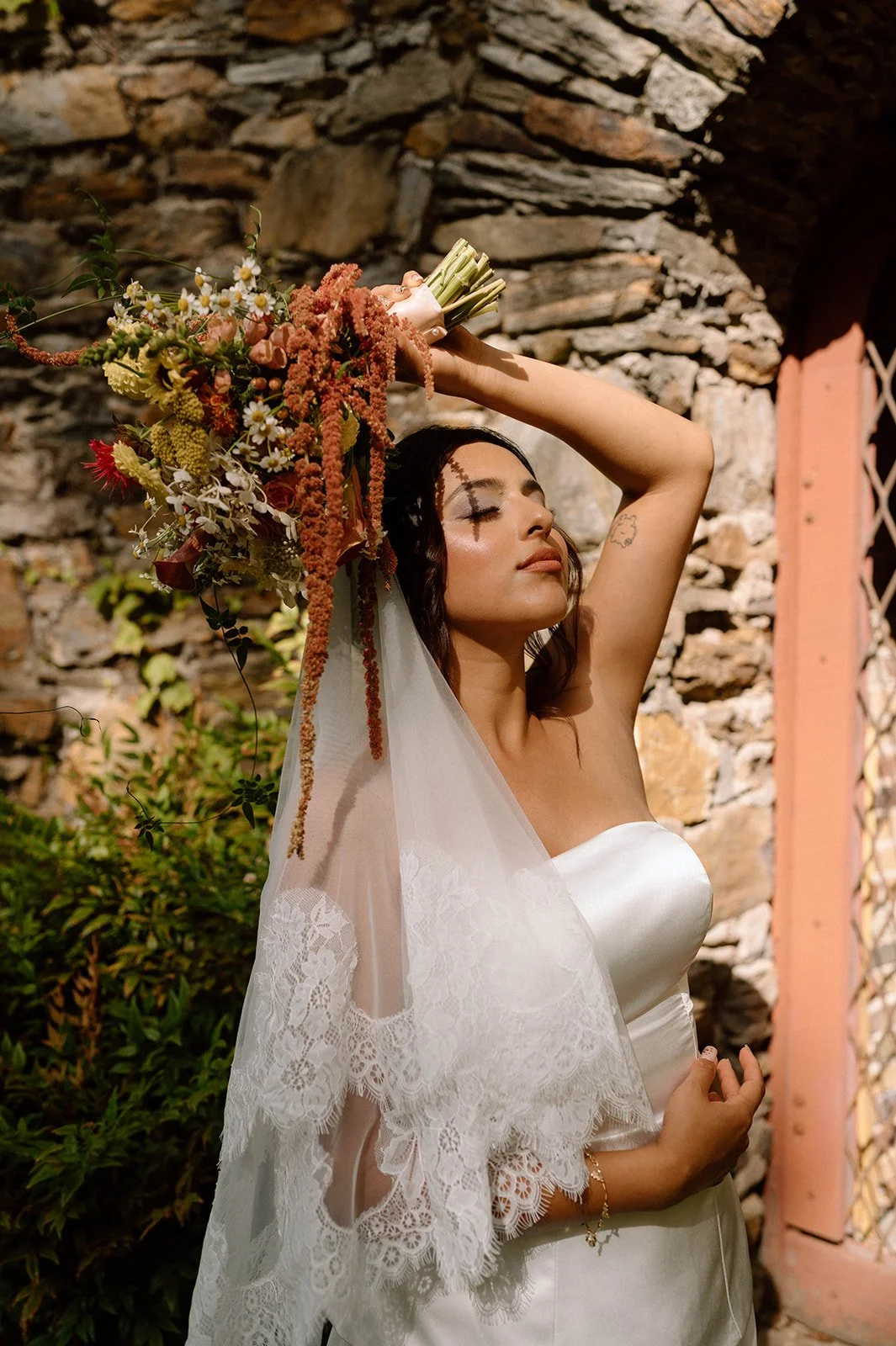 A woman in a white wedding dress holding a bouquet of flowers, standing outdoors with a stone wall in the background.