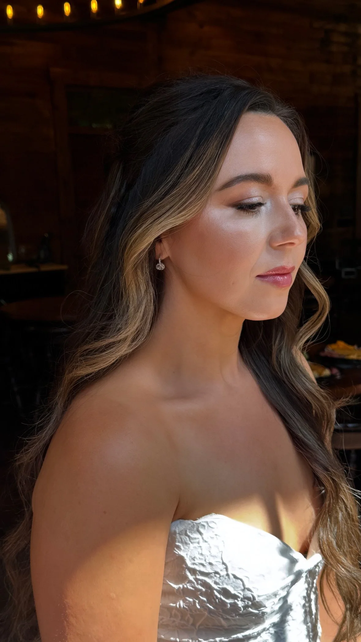 A woman with long, wavy brown hair and light makeup, wearing diamond earrings and a silver strapless dress, standing indoors with warm wooden walls and yellow string lights in the background.