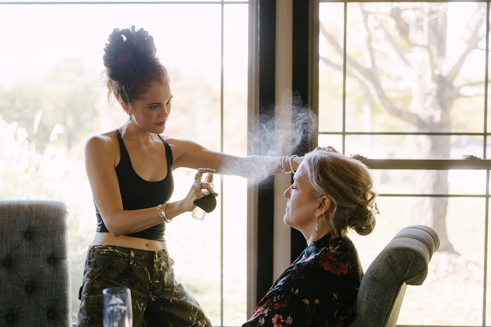 A woman is spraying mist on another woman seated in a chair inside a room with large windows and a view of trees outside.