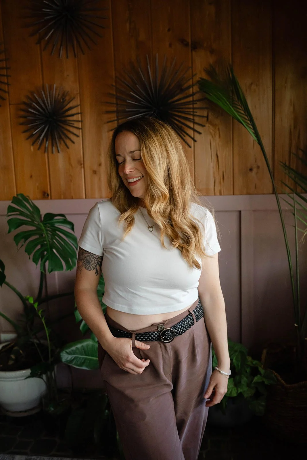 A smiling woman with long wavy hair wearing a white crop top and brown high-waisted pants standing in a room with wooden walls and indoor plants.