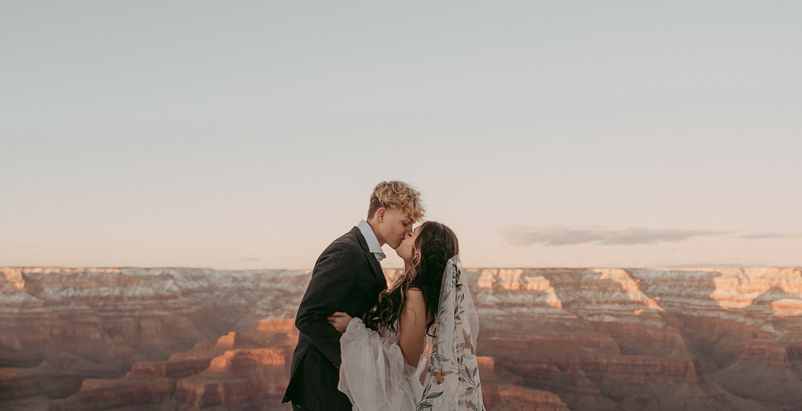 A couple kissing at the Grand Canyon during sunset, with the man in a black suit and the woman in a white dress with a floral veil.