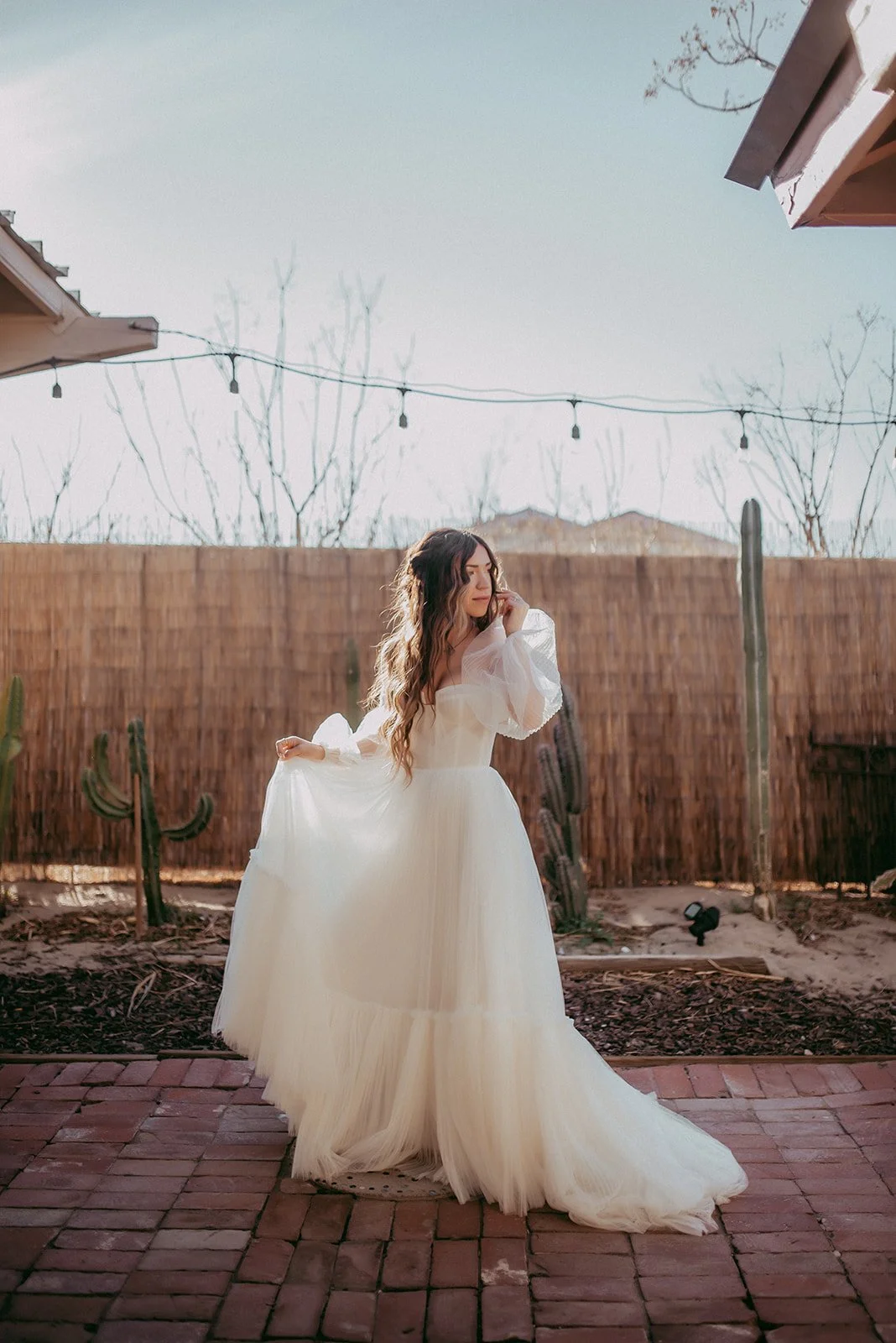 A woman in a white wedding dress standing outdoors on a brick pathway, with a brown wooden fence, cactus plants, and string lights in the background.