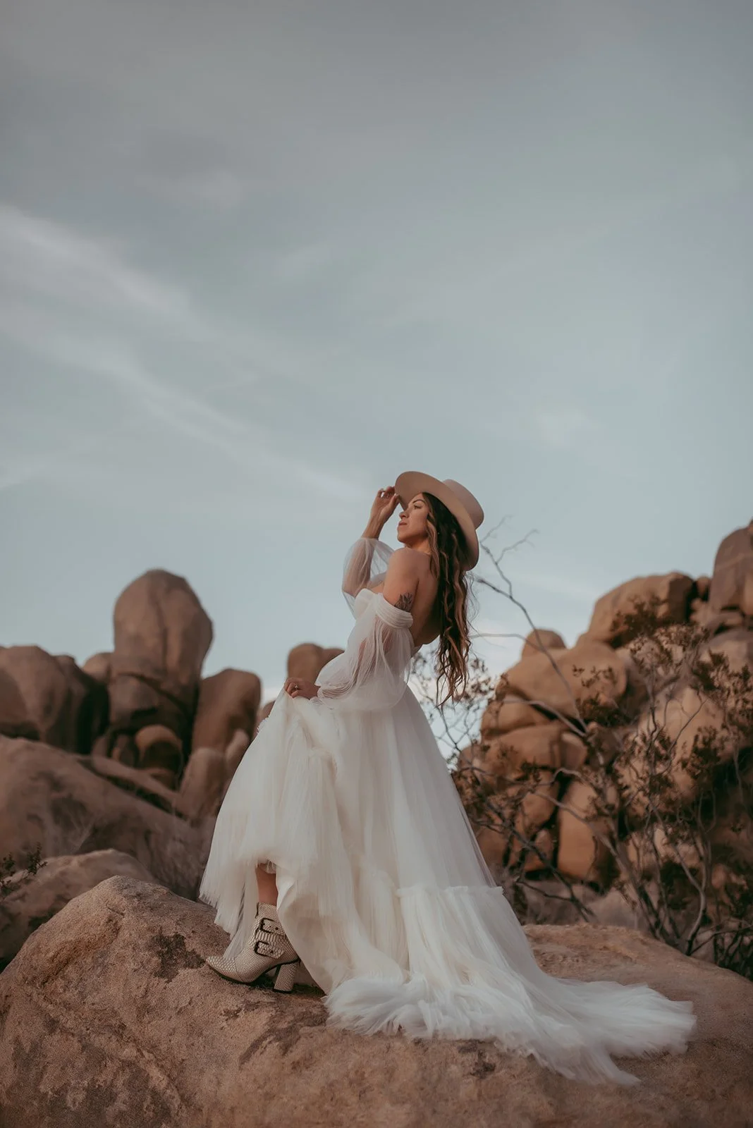 A woman in a white, flowing dress and cowboy boots standing on rocks in a desert landscape, wearing a wide-brim hat.