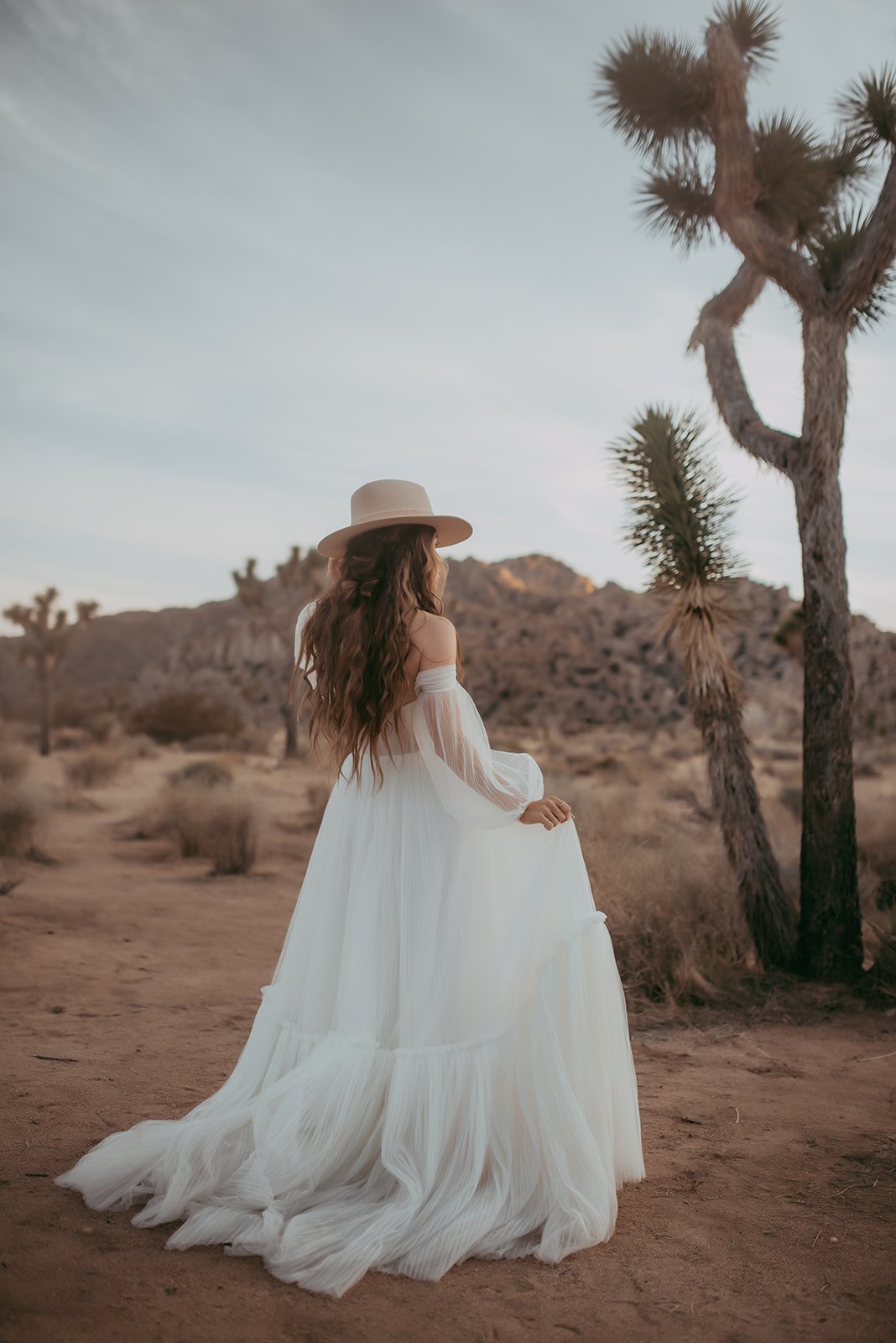 A woman with long wavy hair, wearing a white dress and a wide-brimmed hat, stands in a desert landscape with Joshua trees and rocky hills in the background at sunset.