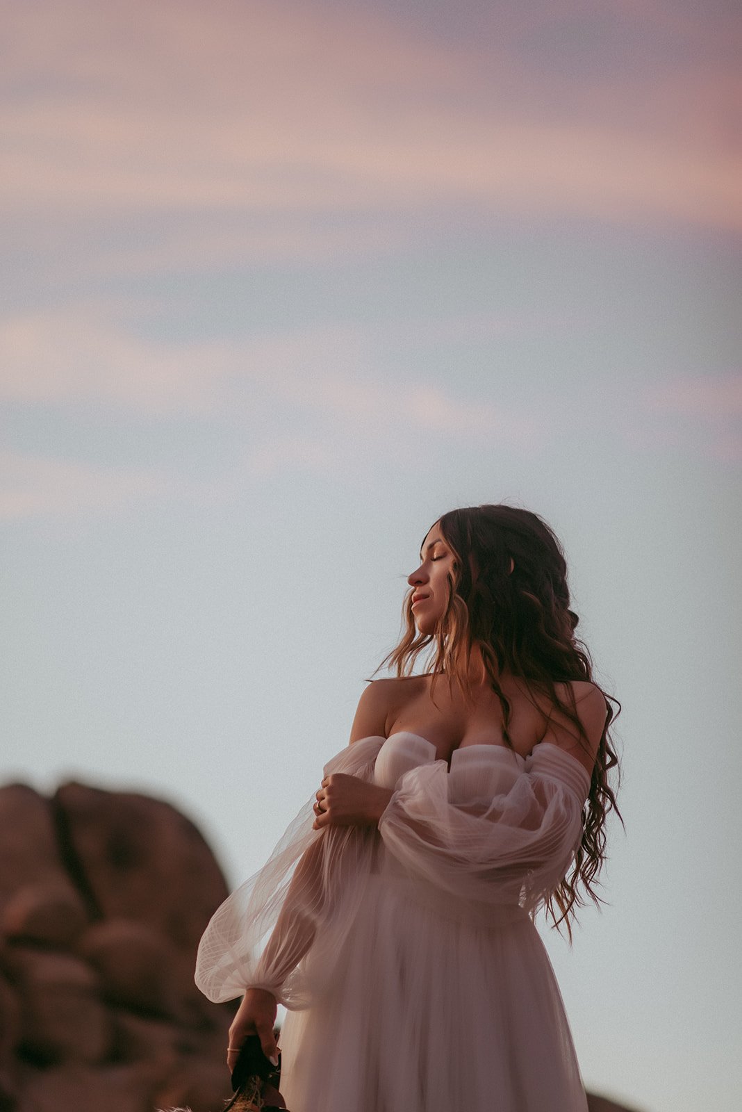 A woman in a flowing off-the-shoulder white dress stands outdoors with arms crossed, eyes closed, during sunset with a pastel-colored sky.