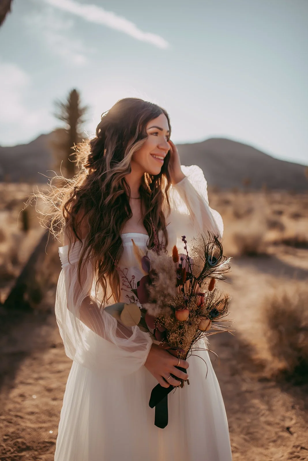 A woman in a white dress smiling and holding a bouquet of flowers outside in a desert landscape with mountains in the background during sunset.