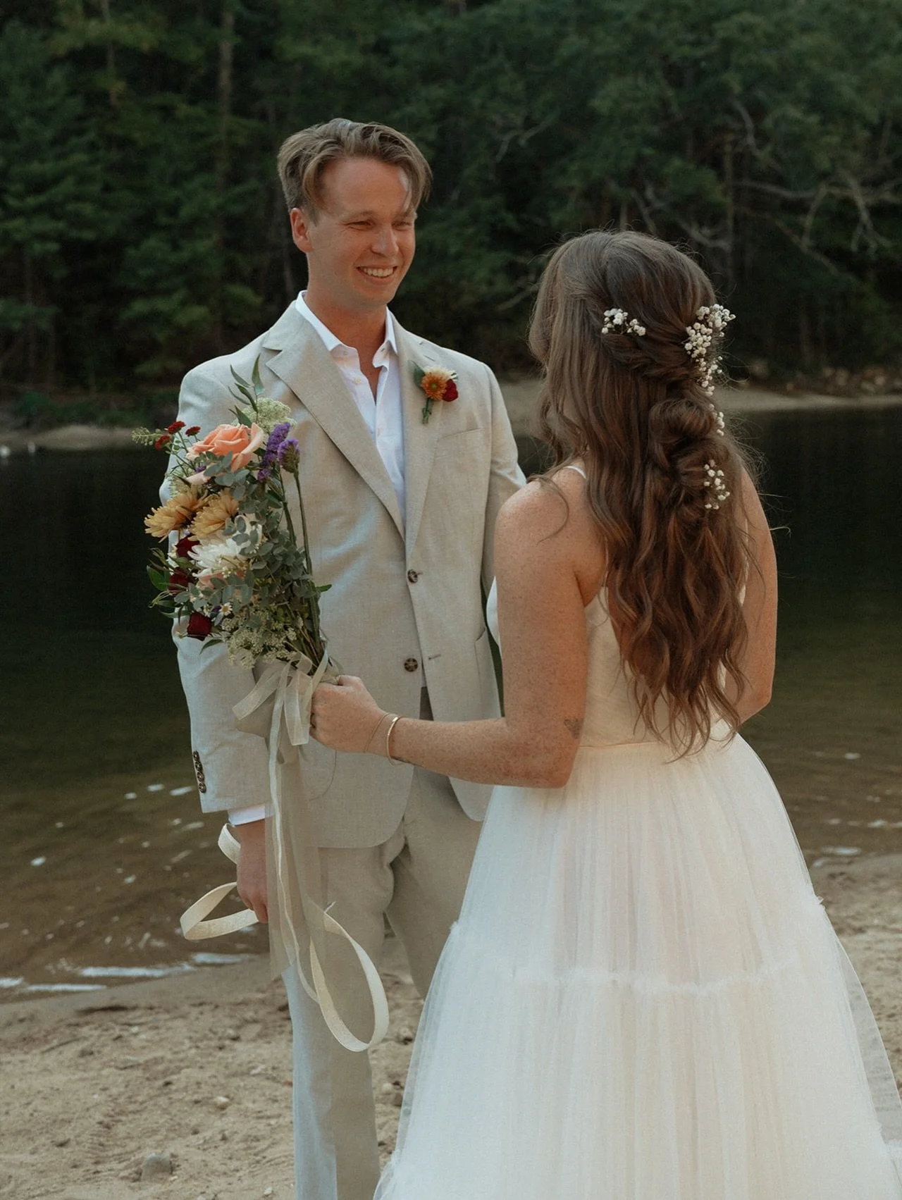 A couple getting married on a beach, with the groom holding a bouquet and the bride with flowers in her hair, standing by water with trees in the background.