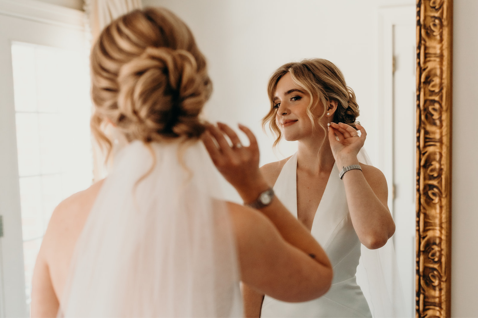 A woman with wavy blonde hair in a loose updo, wearing a white sleeveless dress, is looking at herself in a mirror and adjusting her earring.