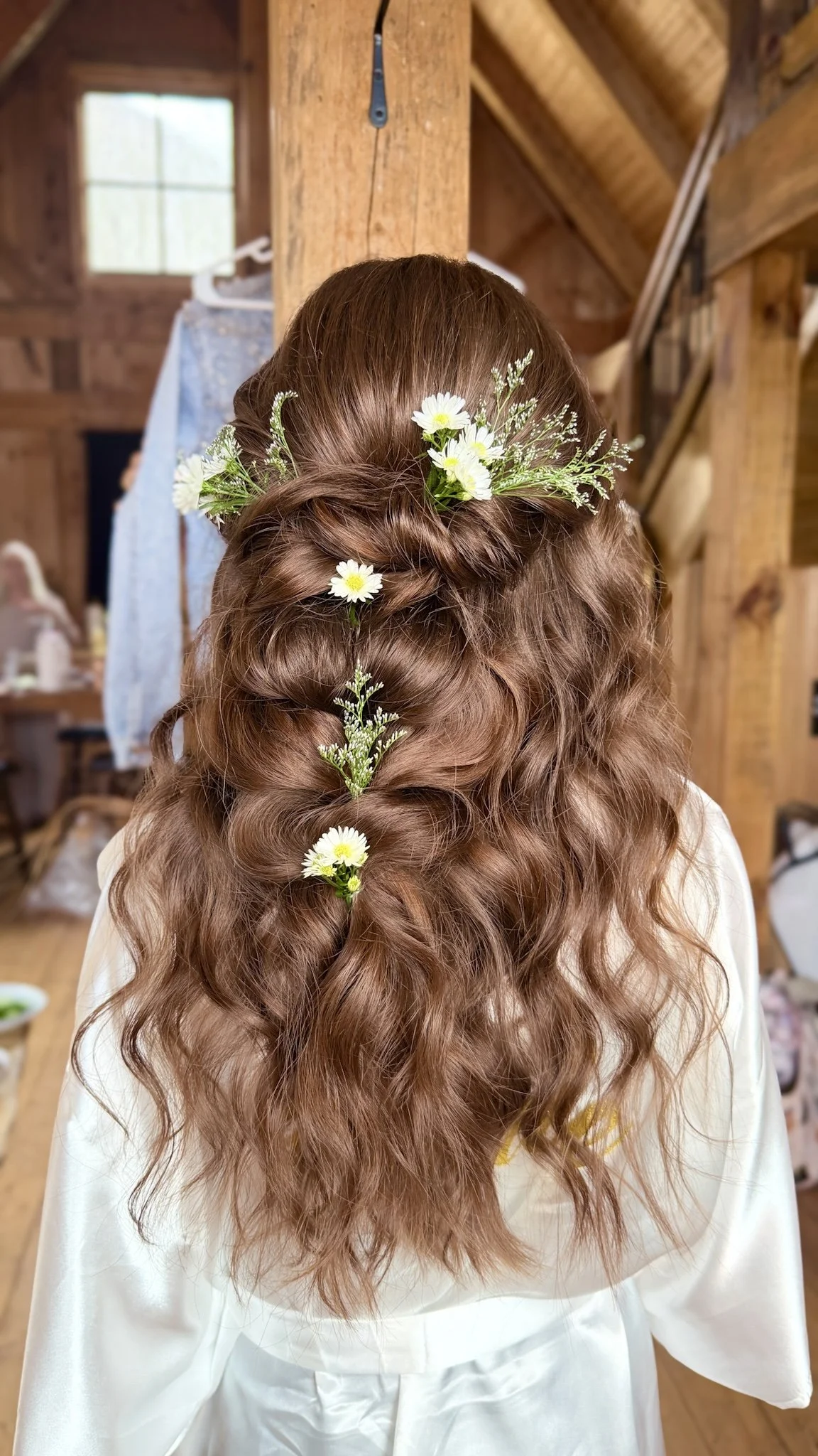 A woman's hairstyle with loose, wavy, auburn hair decorated with white daisies and baby's breath flowers, set in a rustic wooden interior.