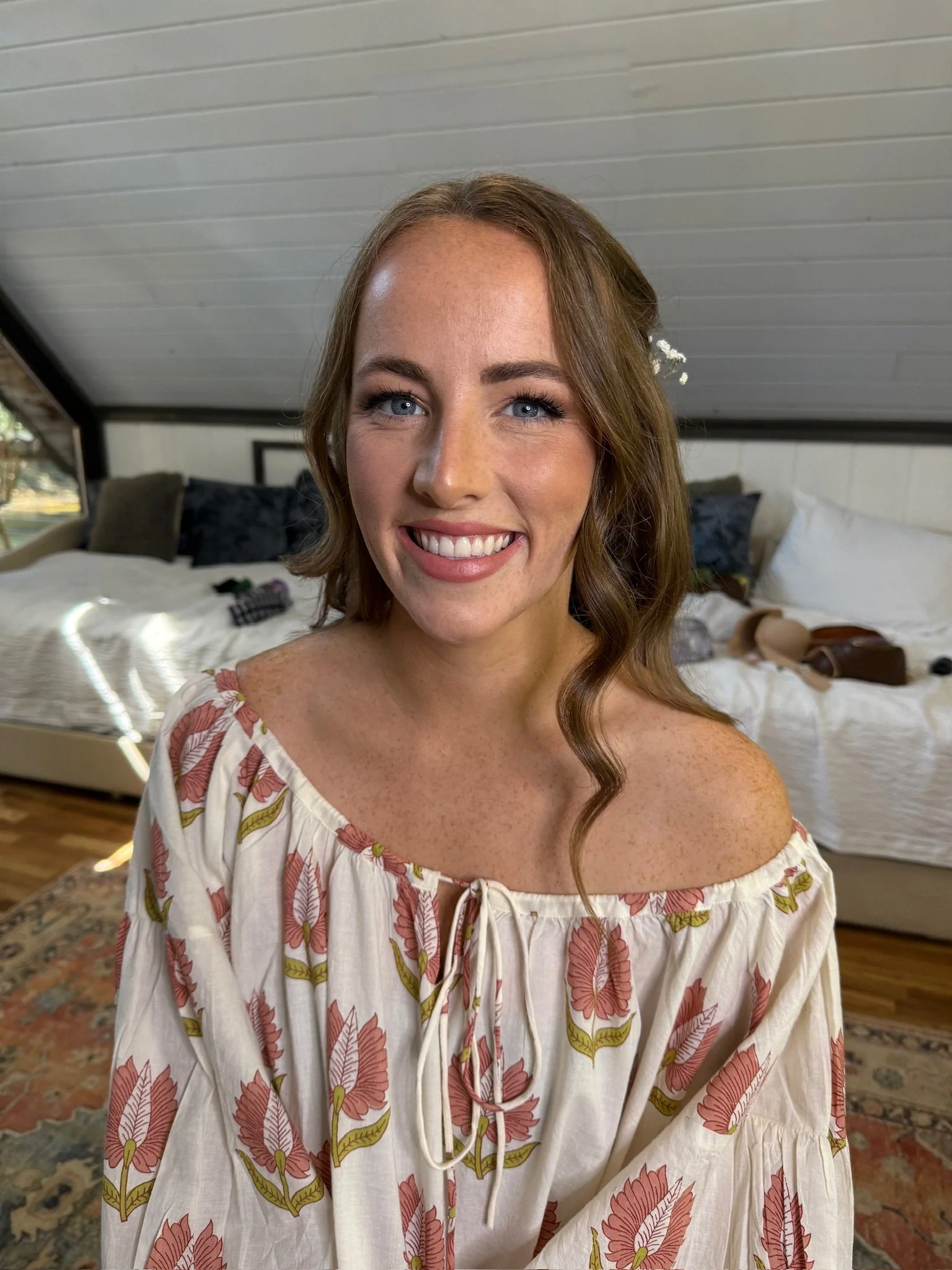 A woman with blue eyes and wavy reddish-brown hair sitting in a room with a bed in the background, wearing a cream-colored off-the-shoulder dress with a pink and green leaf pattern, and a small white flower in her hair.