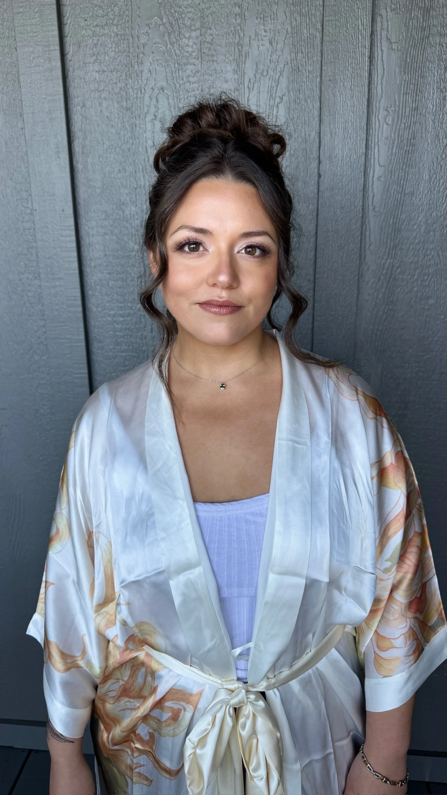 A woman with light skin and dark brown hair styled in an updo with loose curls framing her face, wearing a satin robe with a floral pattern and a white tank top underneath, standing against a grey textured wall.