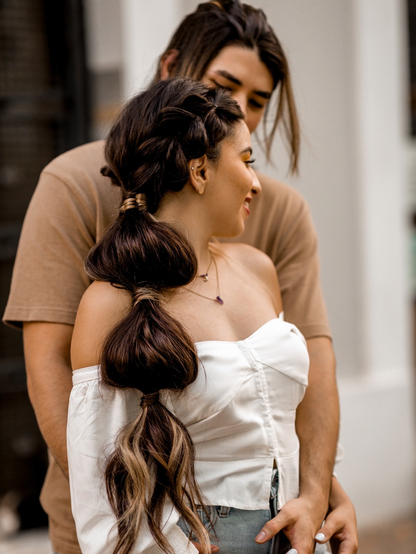 Two women with long, dark, curled hair sharing an intimate moment, with one woman standing behind the other and resting her head on the other's forehead.