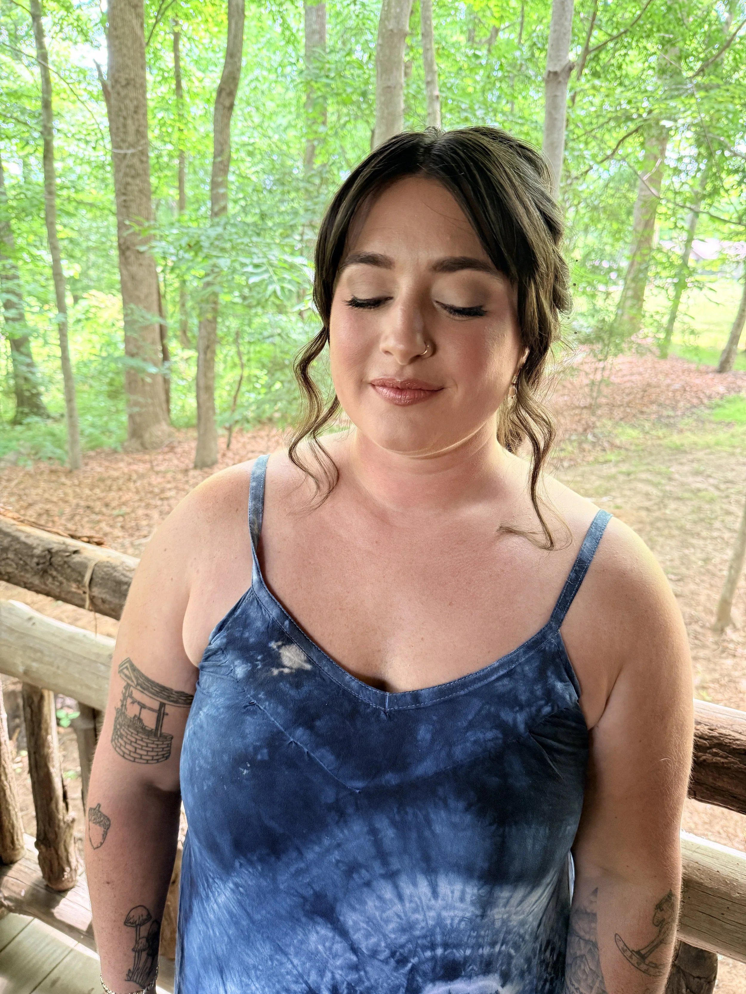 A woman with short dark hair with waves, closed eyes, and a nose ring, wearing a blue tie-dye sleeveless top, standing outdoors on a wooden bridge in a forest with green leaves and tree trunks in the background.