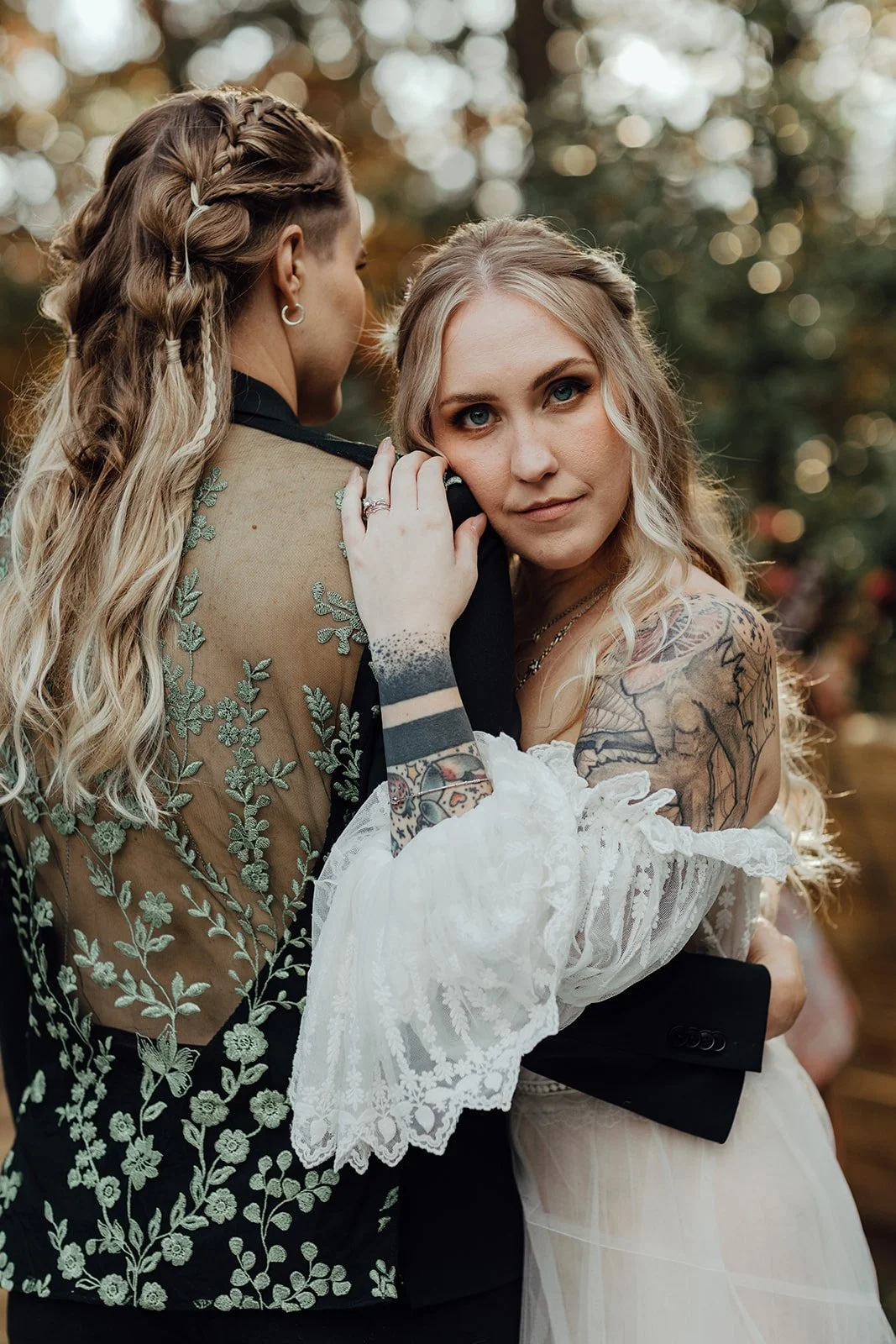 Two women, one with long blonde hair and tattoos, and the other with curly blonde hair in a braid, embrace outdoors with blurred trees in the background.