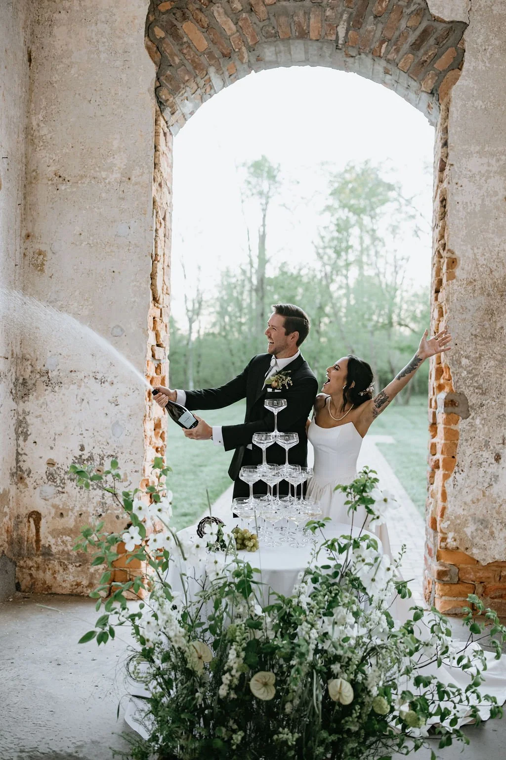 A newlywed couple celebrating a wedding, with the groom spraying champagne and the bride smiling and raising her arm. They are standing behind a table with a pyramid of empty glasses and floral decorations, in a rustic setting with a large arched window and outdoor greenery in the background.