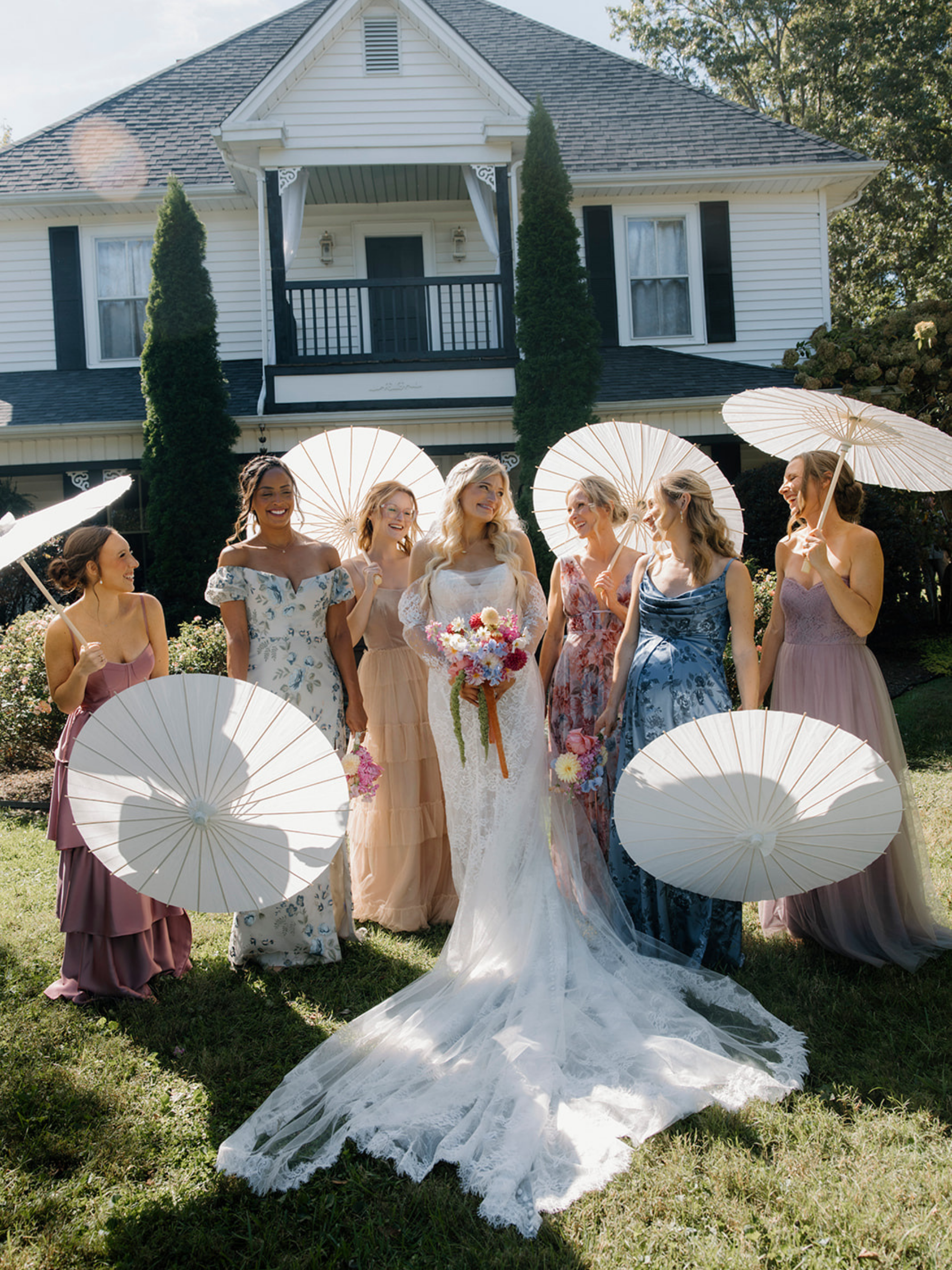 A bride in a white wedding dress holding a bouquet, surrounded by women in colorful dresses holding white parasols, standing on a lawn in front of a house with white siding and black shutters.