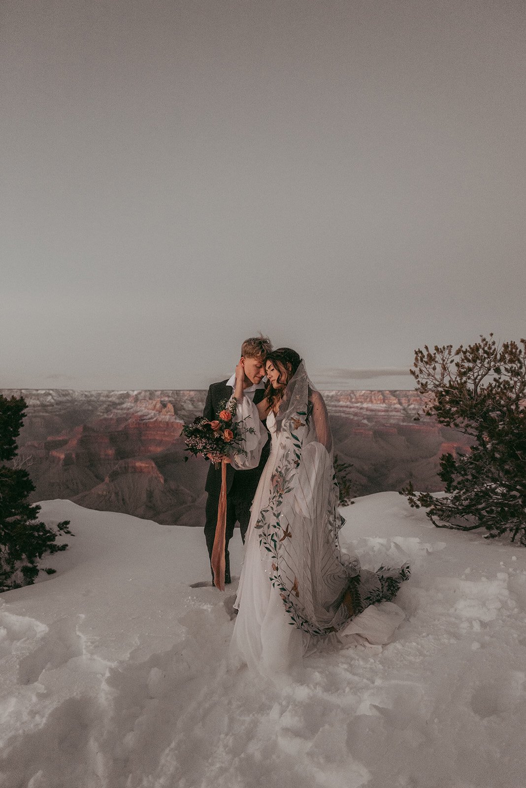 A bride and groom embrace on snow-covered ground overlooking the Grand Canyon at sunset, with trees framing the scene.