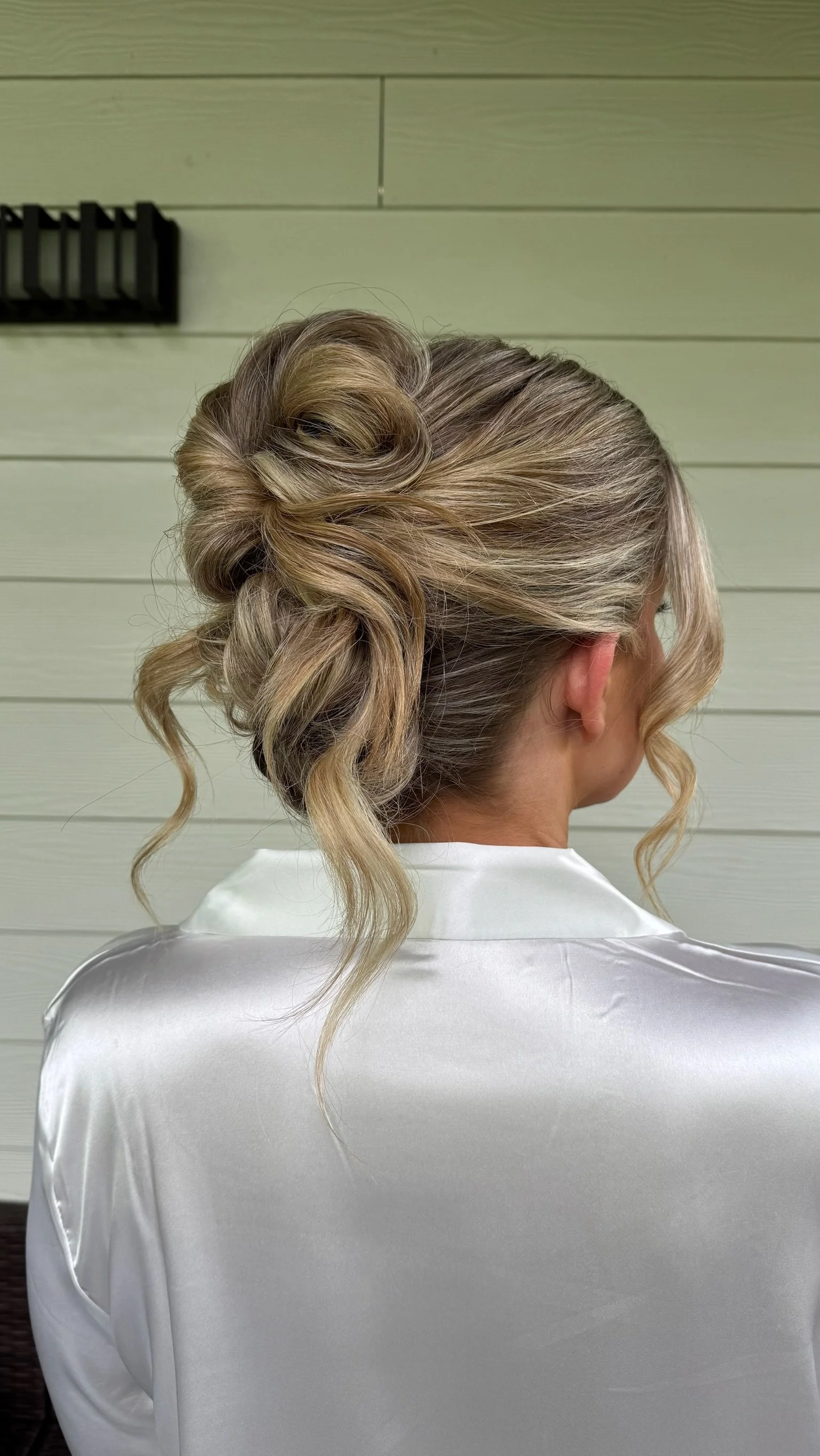 A woman with styled blonde hair and loose curls, wearing a white satin robe, seen from behind against a light-colored wooden wall.