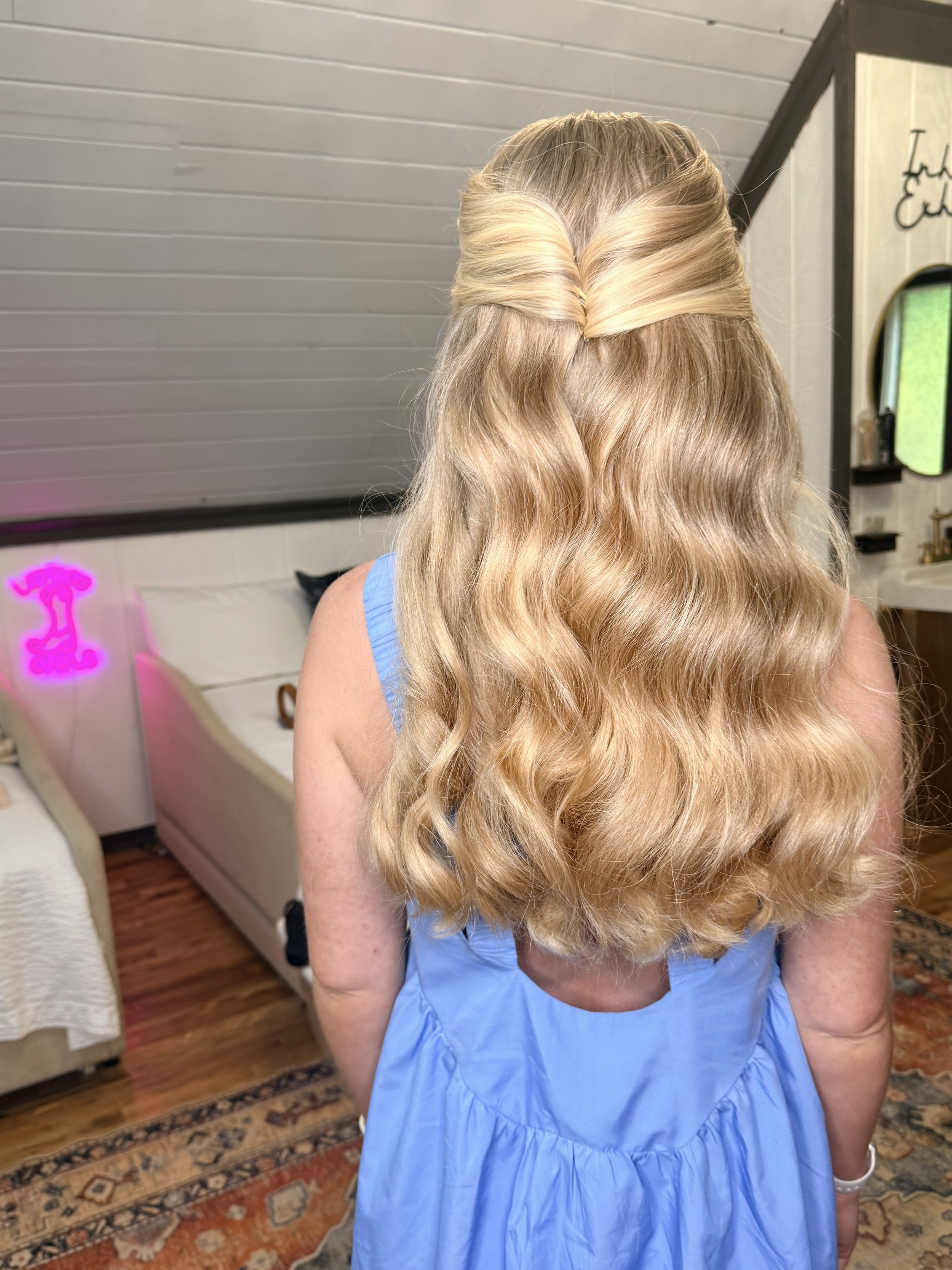 Back view of a woman with long, wavy blonde hair falling over her shoulders, wearing a blue dress or gown inside a room with wooden flooring and a patterned rug.