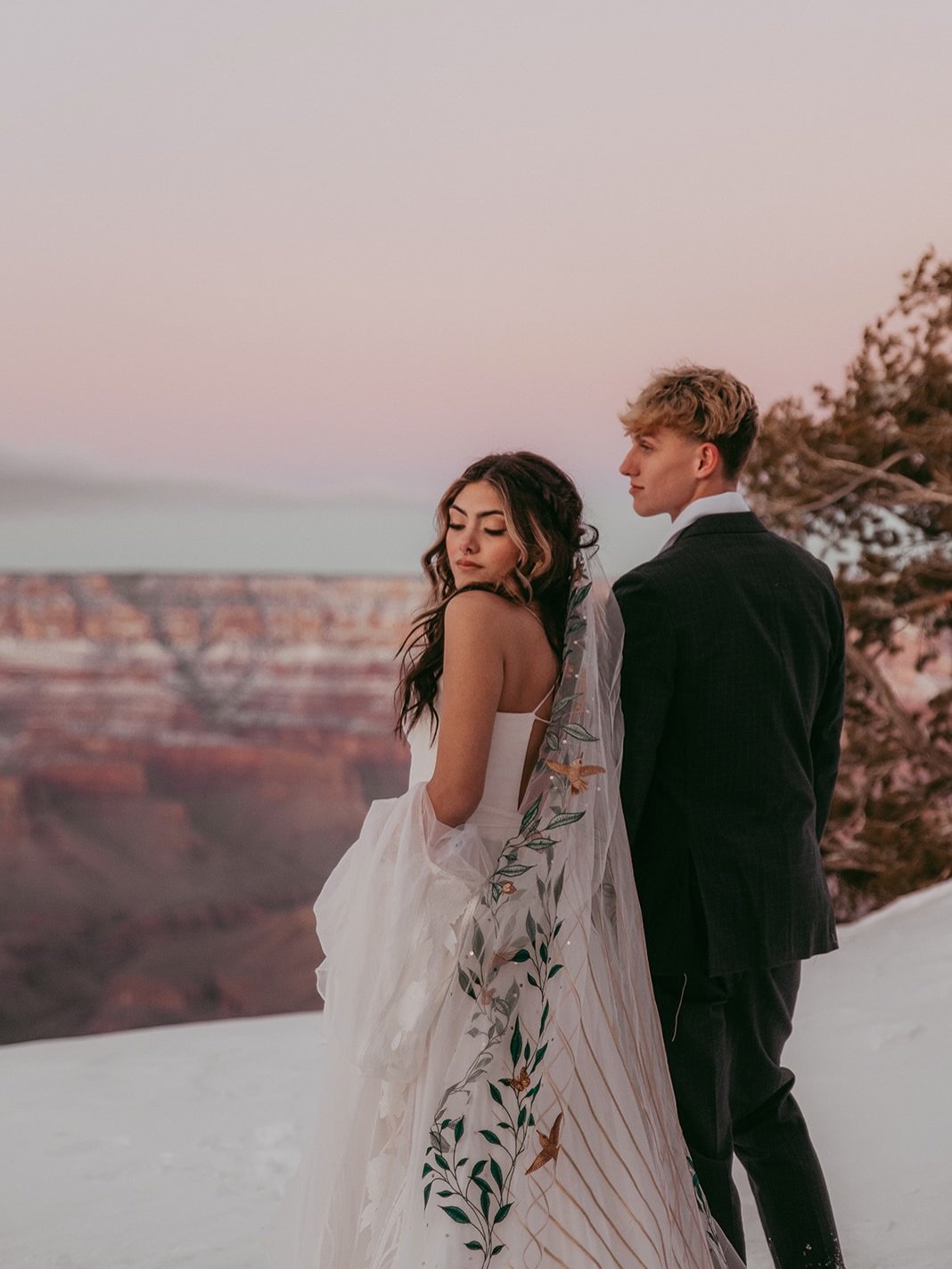 A couple dressed in wedding attire posing on a snowy landscape at sunset with canyon views in the background.