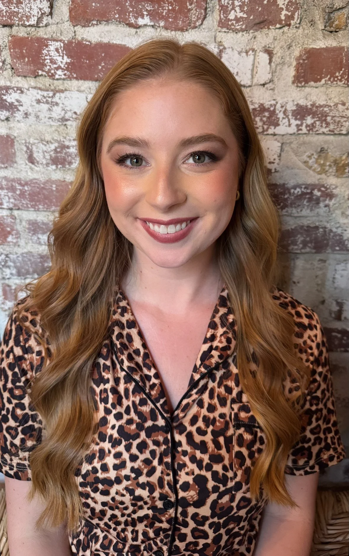 A woman with long wavy reddish-brown hair smiling in front of a brick wall, wearing a leopard print blouse.