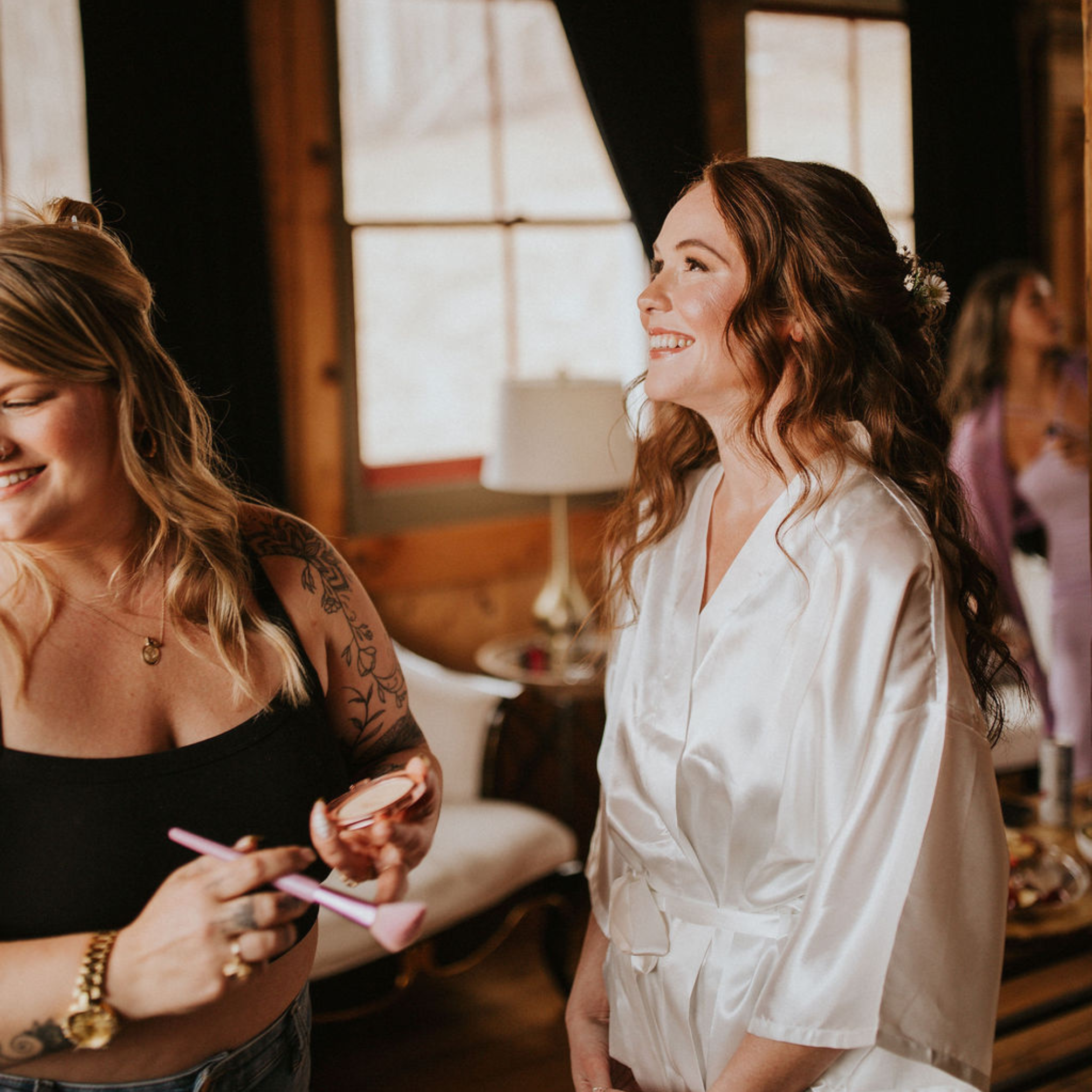 Two women are standing close together, with one woman showing something on a pink phone to the other woman. The woman on the left has tattoos on her arm, a bracelet, and is smiling with her eyes closed. The woman on the right has long brown hair and is smiling while looking at the phone. They are indoors near a window and wooden walls.