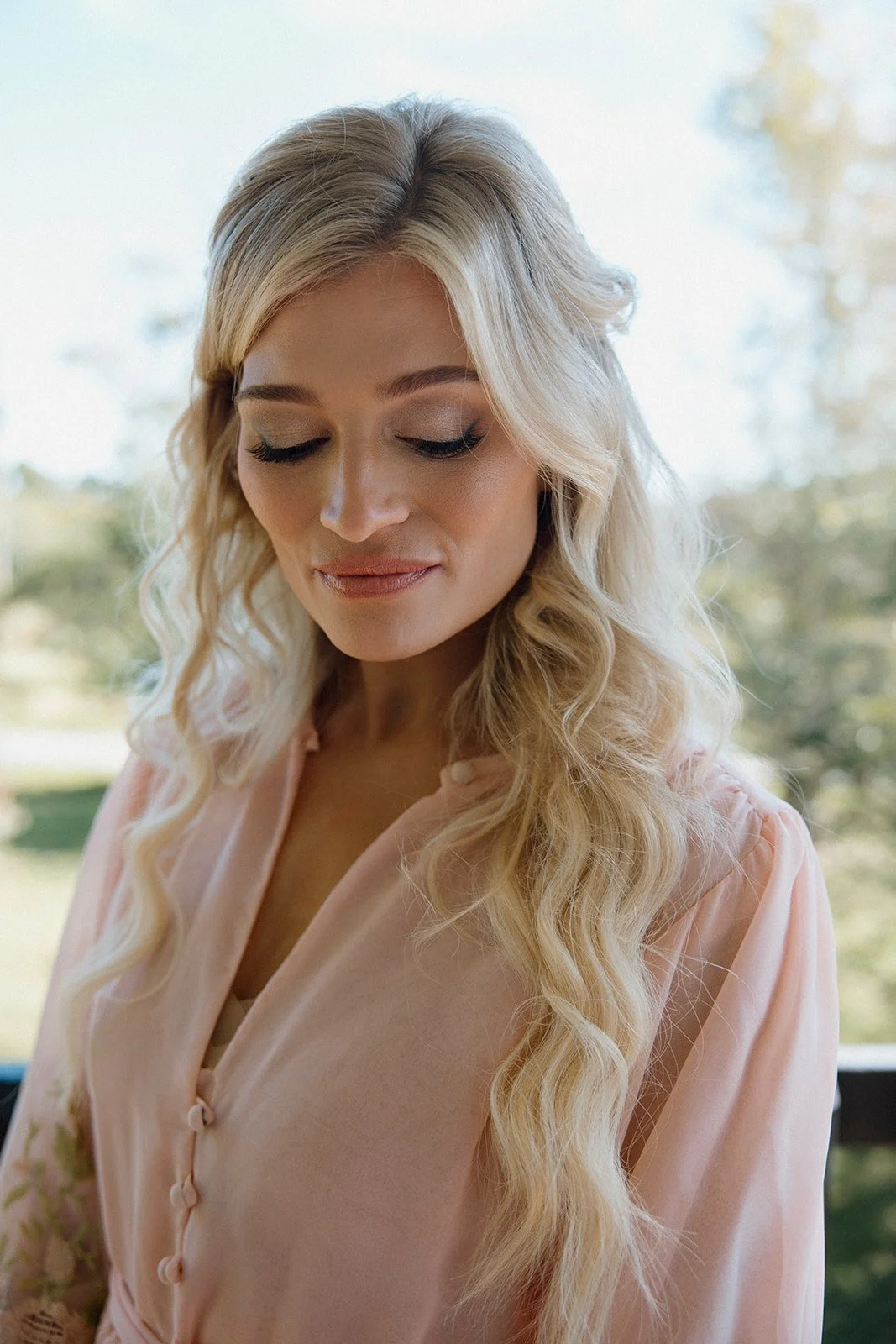Close-up of a woman with long, curly blonde hair and closed eyes, wearing a peach-colored blouse with decorative buttons, standing outdoors with a blurred background of trees and sky.