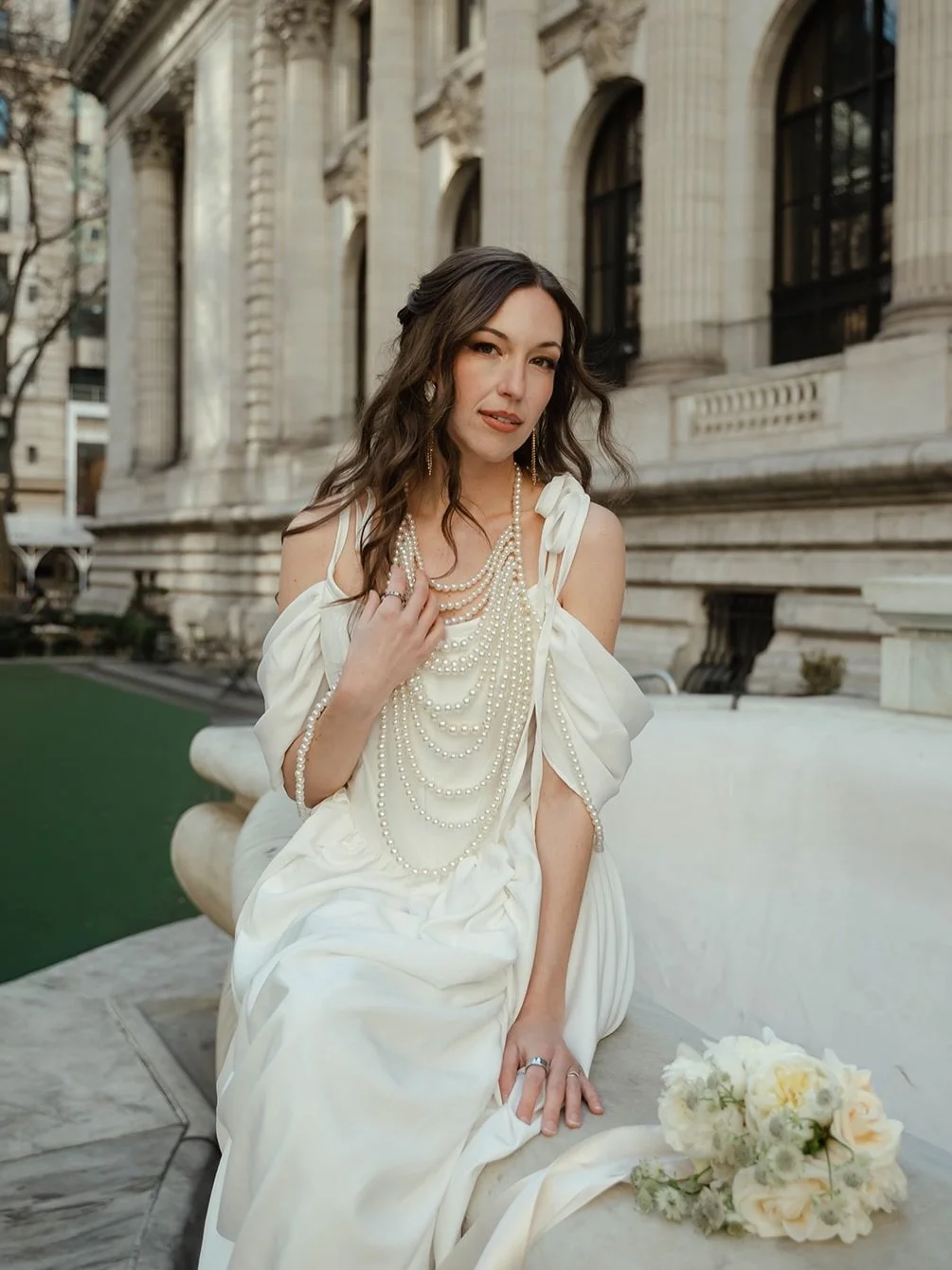 Woman sitting on a marble bench in front of a historic building dressed in a white gown with pearl necklaces and earrings, holding a bouquet of white flowers.