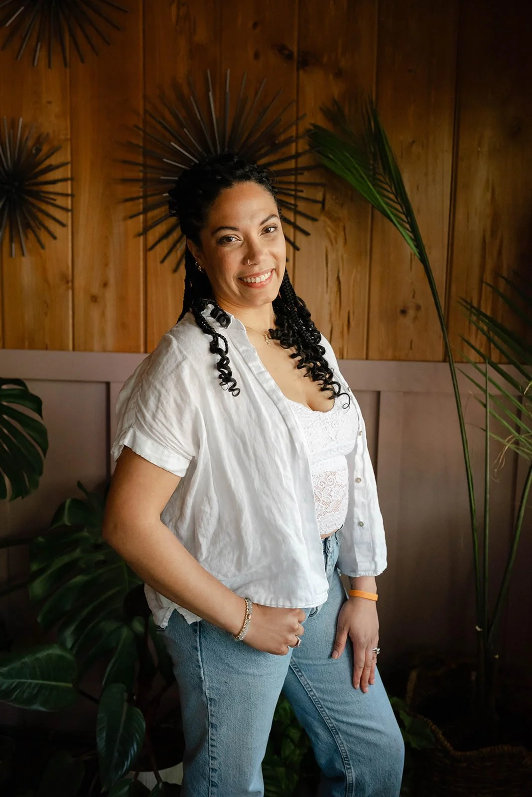 A woman with braided black hair smiling, standing indoors with a wooden wall decorated with metal wall art and surrounded by green plants.