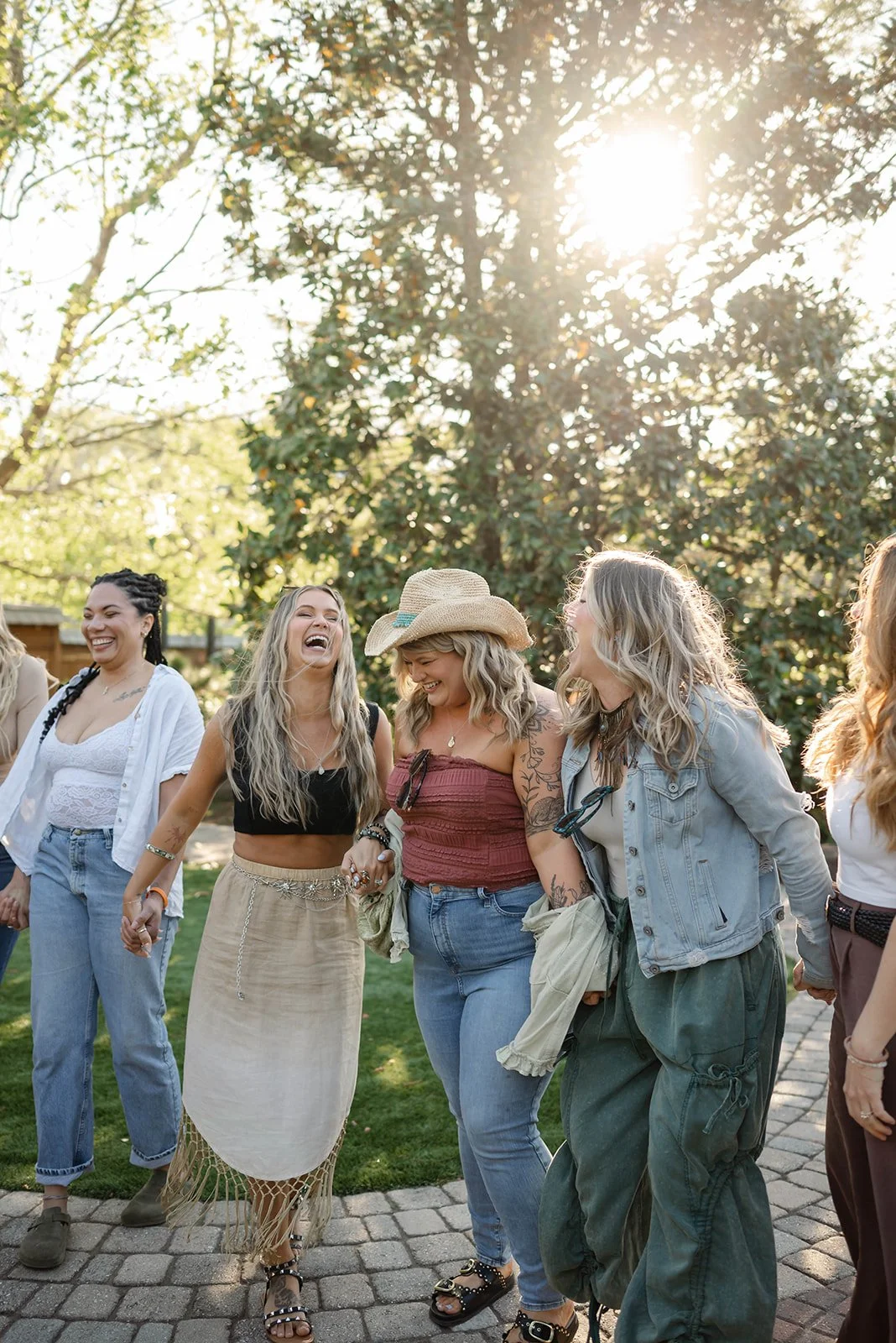 A group of women holding hands, laughing, and enjoying a sunny day outdoors in a park or garden setting.