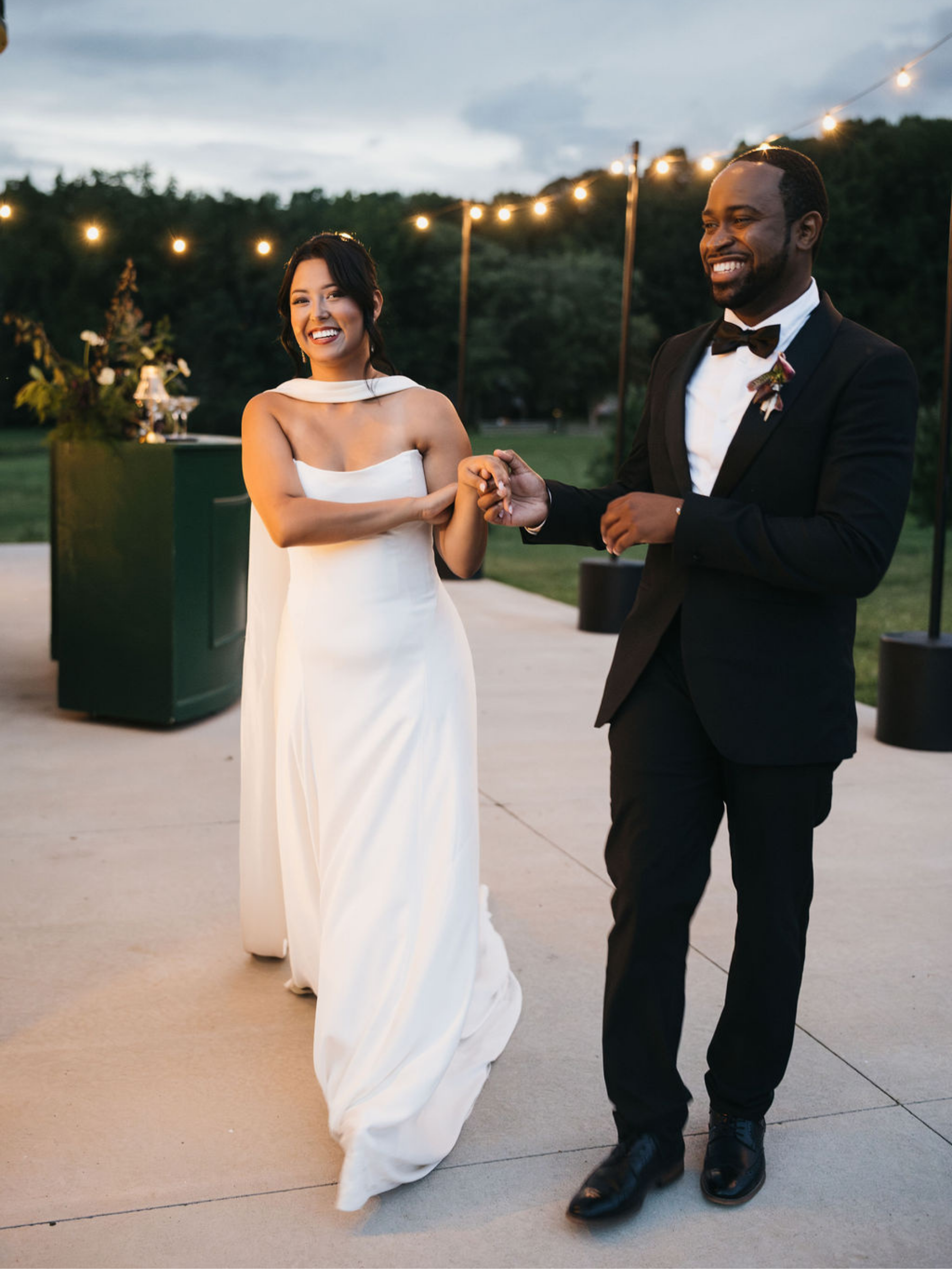 A newlywed couple smiling and dancing outdoors during the evening, with string lights overhead and a lush green background.