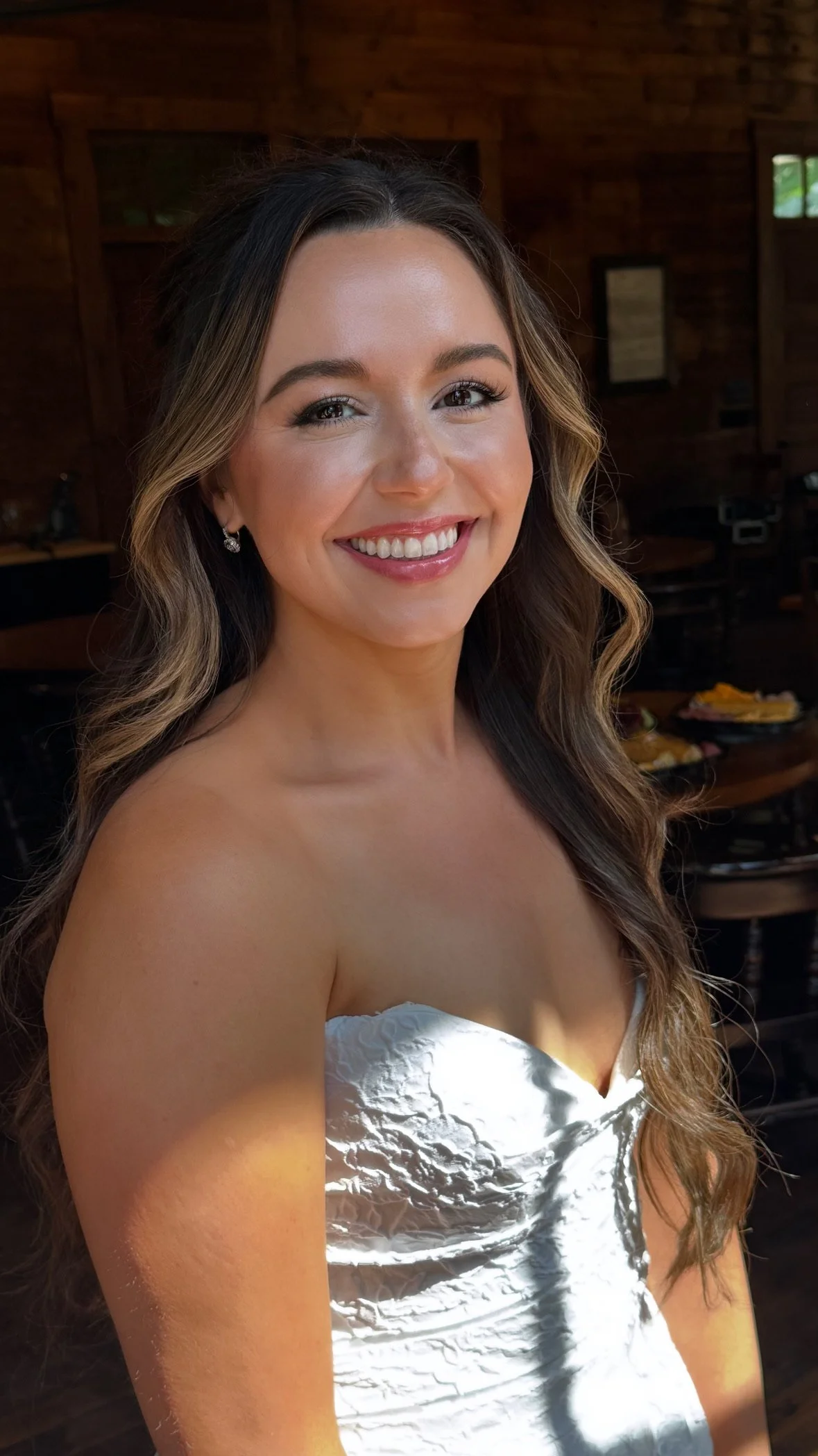 A woman smiling with long, wavy brown hair, wearing a strapless silver dress, in a rustic wooden interior.