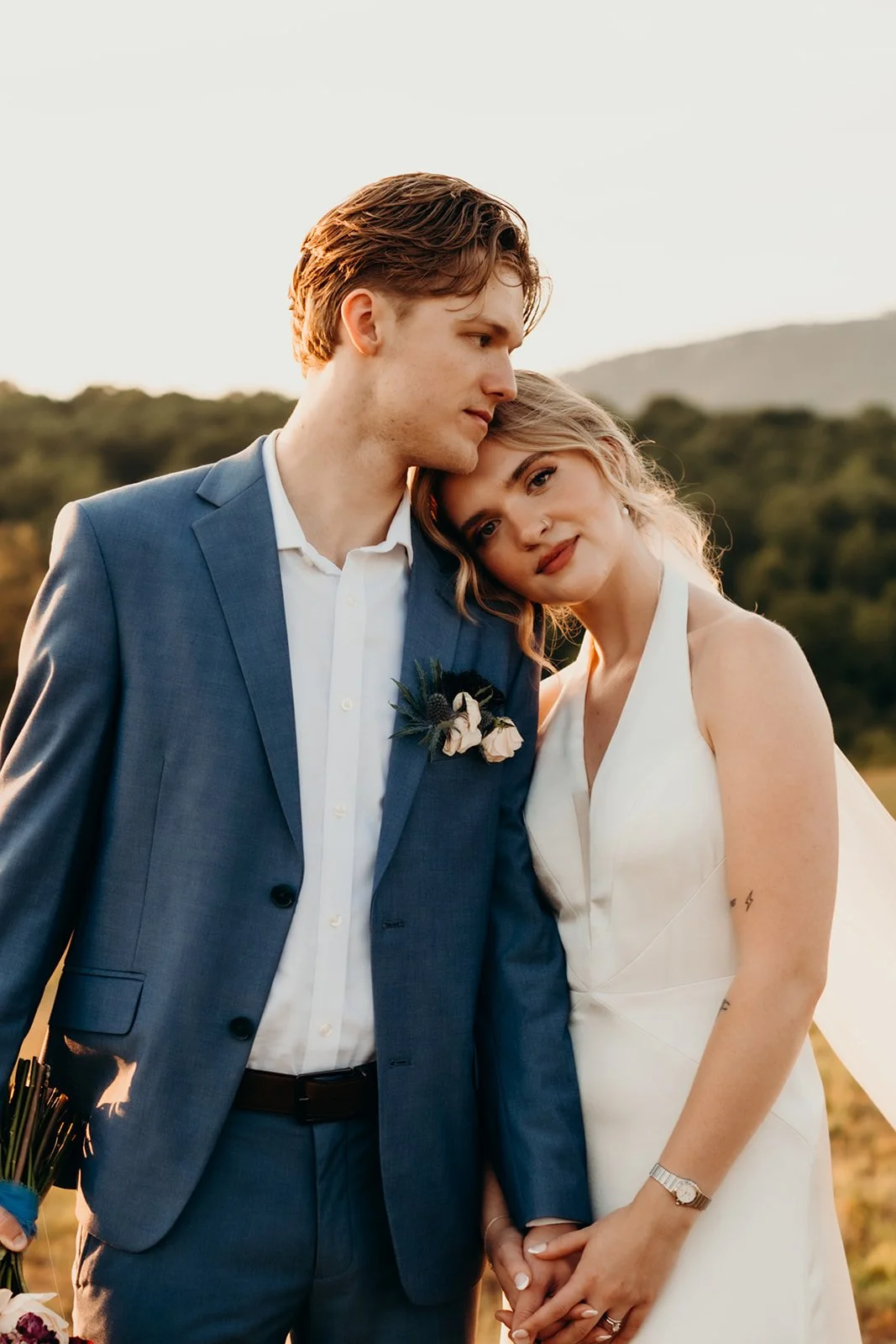 A couple dressed in wedding attire, standing close together outdoors, with the woman leaning her head on the man's shoulder, both smiling softly. The man is wearing a blue suit with a white shirt, and the woman is in a white wedding dress.