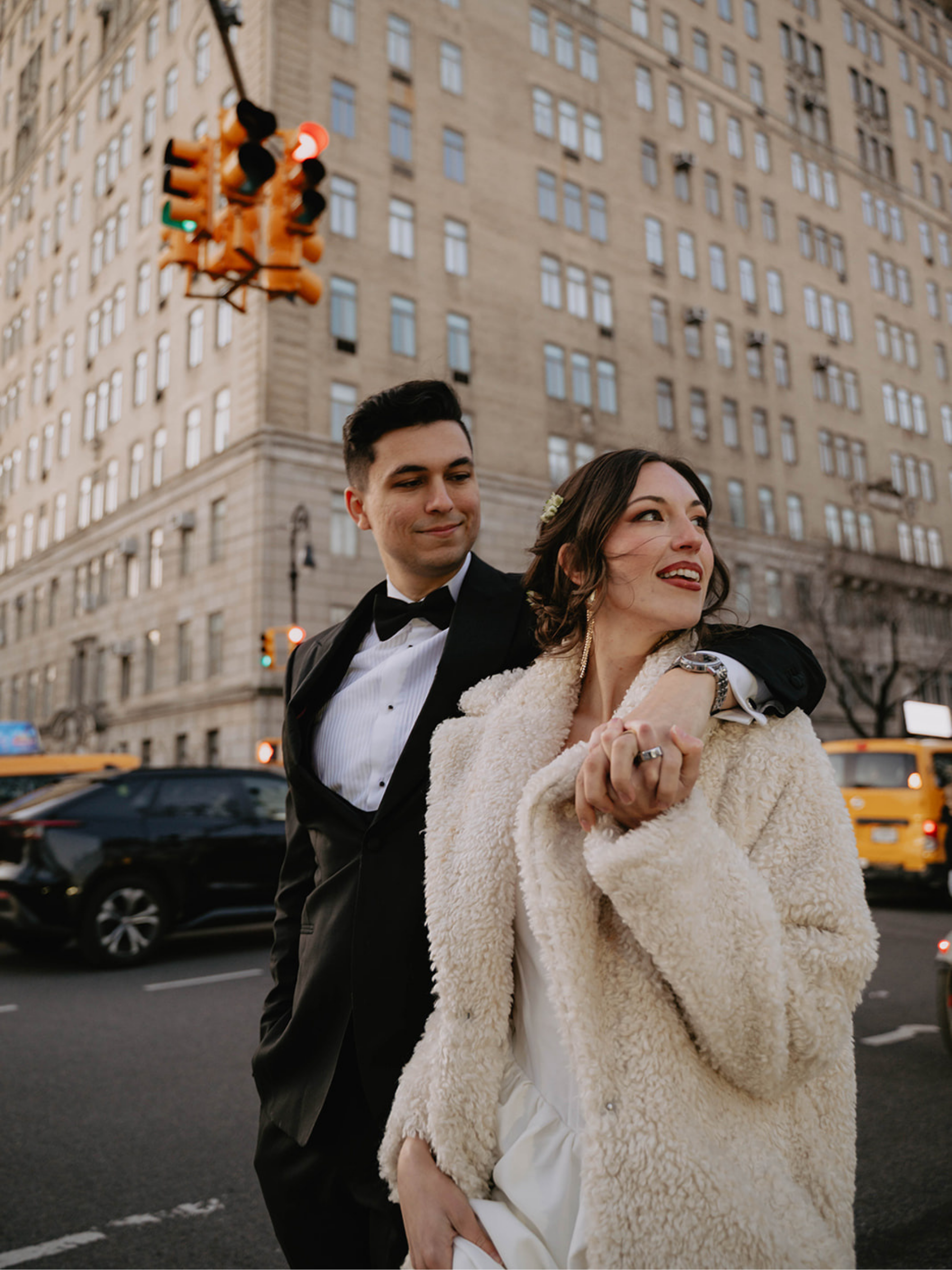 A young couple in formal attire standing in the city at dusk, with tall buildings in the background and traffic lights overhead.