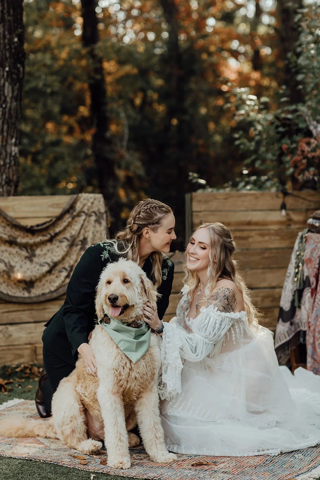 Two women, one in a wedding dress and the other in a black suit, smile at each other in an outdoor setting with a large dog. The woman in the dress has long blonde hair and tattoos, and the woman in the suit has braided hair. The dog is a large, cream-colored furry breed wearing a green bandana, sitting on a colorful rug in front of a wooden fence with autumn trees in the background.